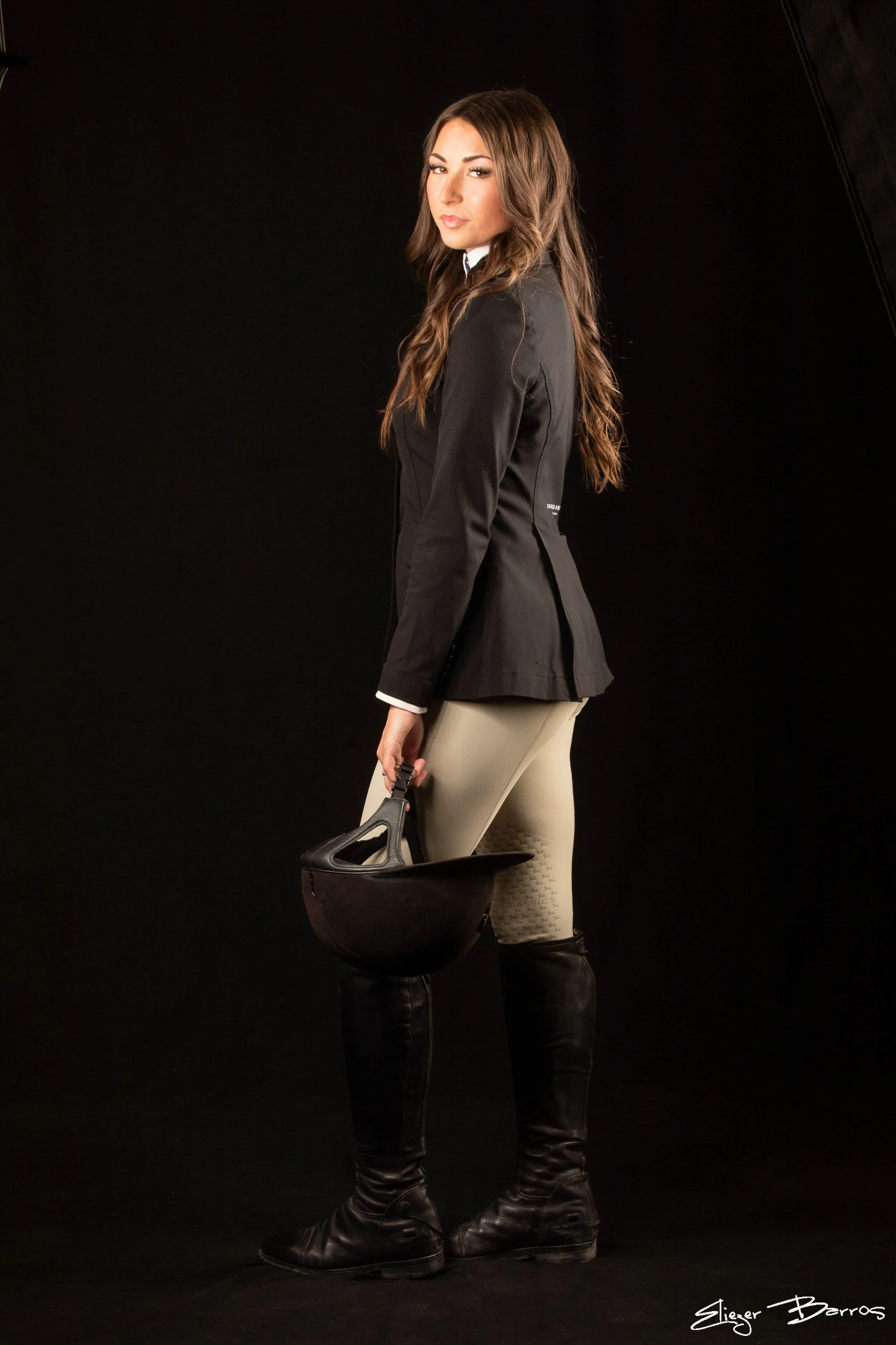 A woman with long brown hair wearing a black blazer, beige riding pants, and black riding boots, holding a black riding helmet in her right hand, standing against a dark background.