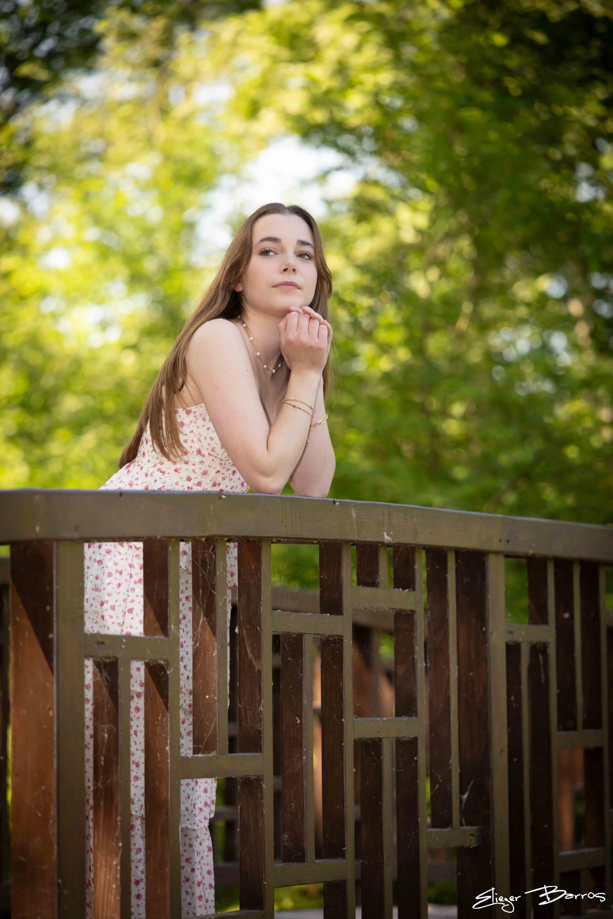 A woman with long brown hair and light skin, wearing a pink floral dress and jewelry, is leaning on a wooden fence with a backdrop of green, leafy trees, softly lit by natural sunlight.