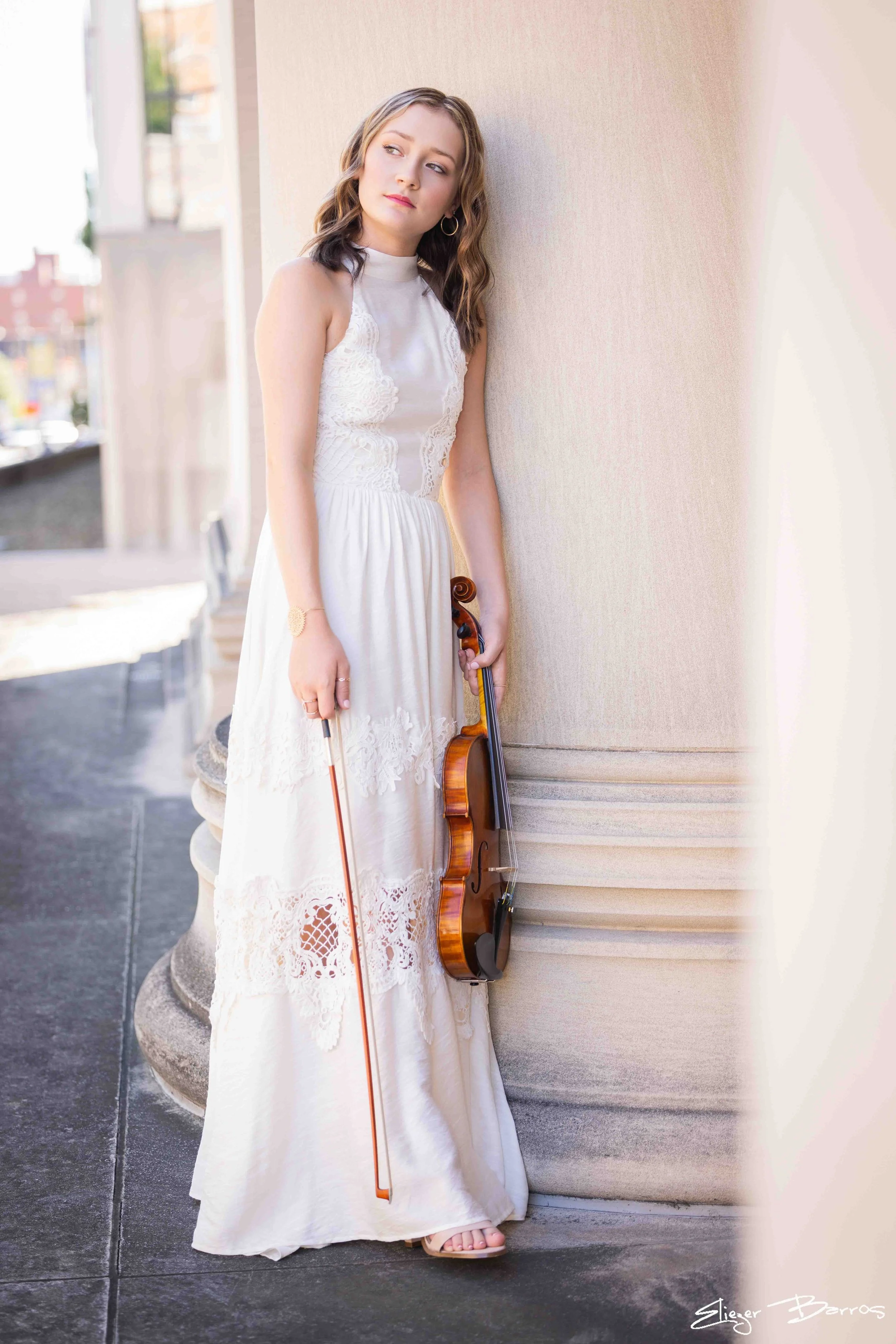 A young woman in a white lace dress is standing outdoors against a beige wall with a violin in her right hand, resting on the ground. She has wavy brown hair and is looking to her left.