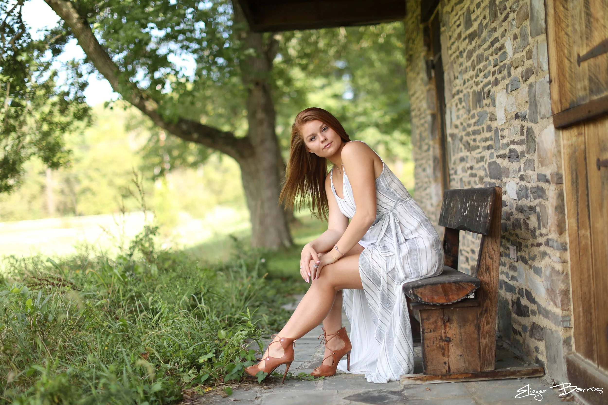 A young woman with long red hair sitting on a wooden bench outdoors beside a stone wall, wearing a white striped dress and brown high heels, looking at the camera with a slight smile, surrounded by green foliage and trees.