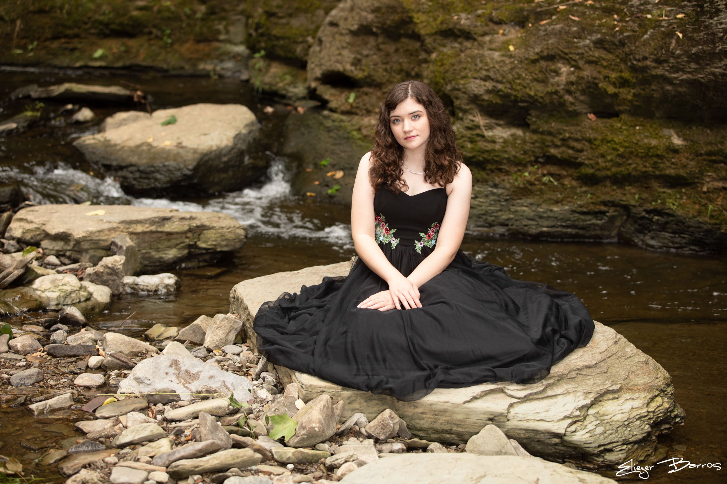 A young woman with curly brown hair sitting by a creek in a wooded area, wearing a black dress with floral embroidery.