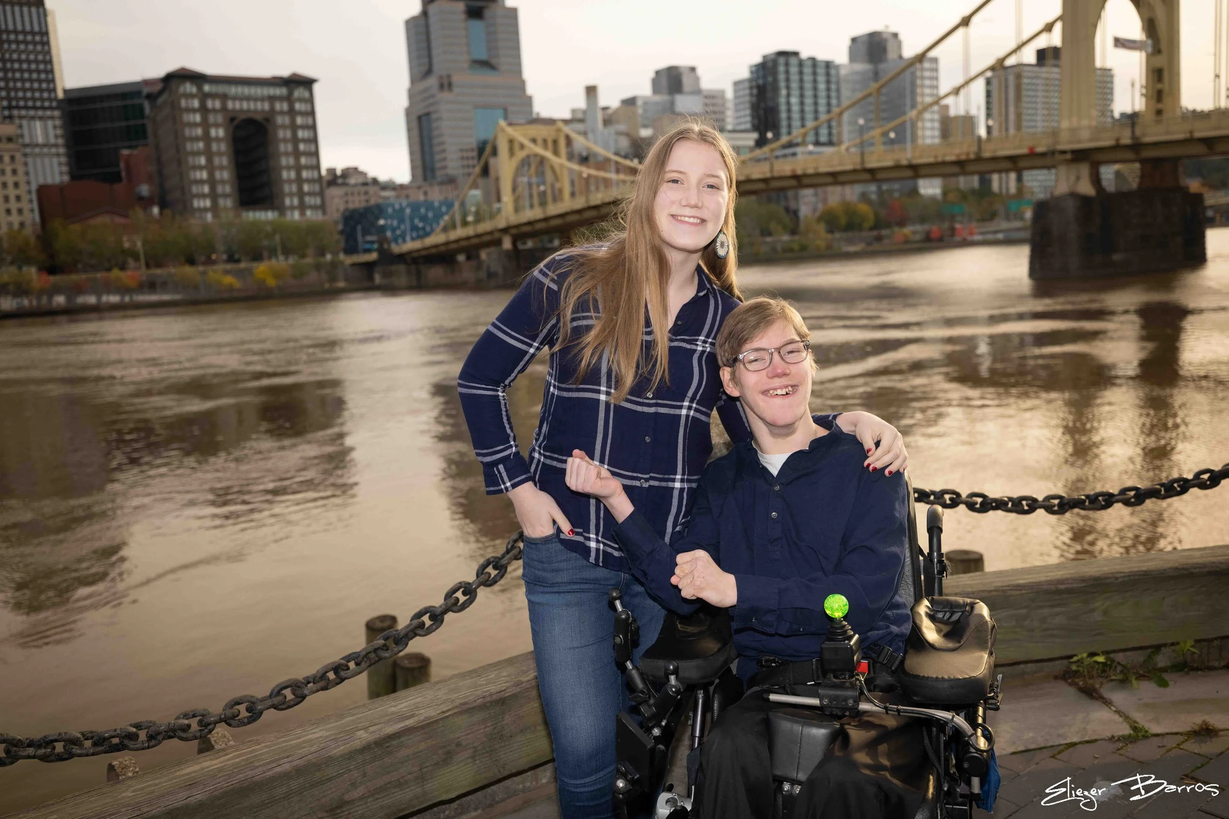 Two smiling girls, one standing and one seated in a wheelchair, by a river with a city skyline and a yellow bridge in the background.
