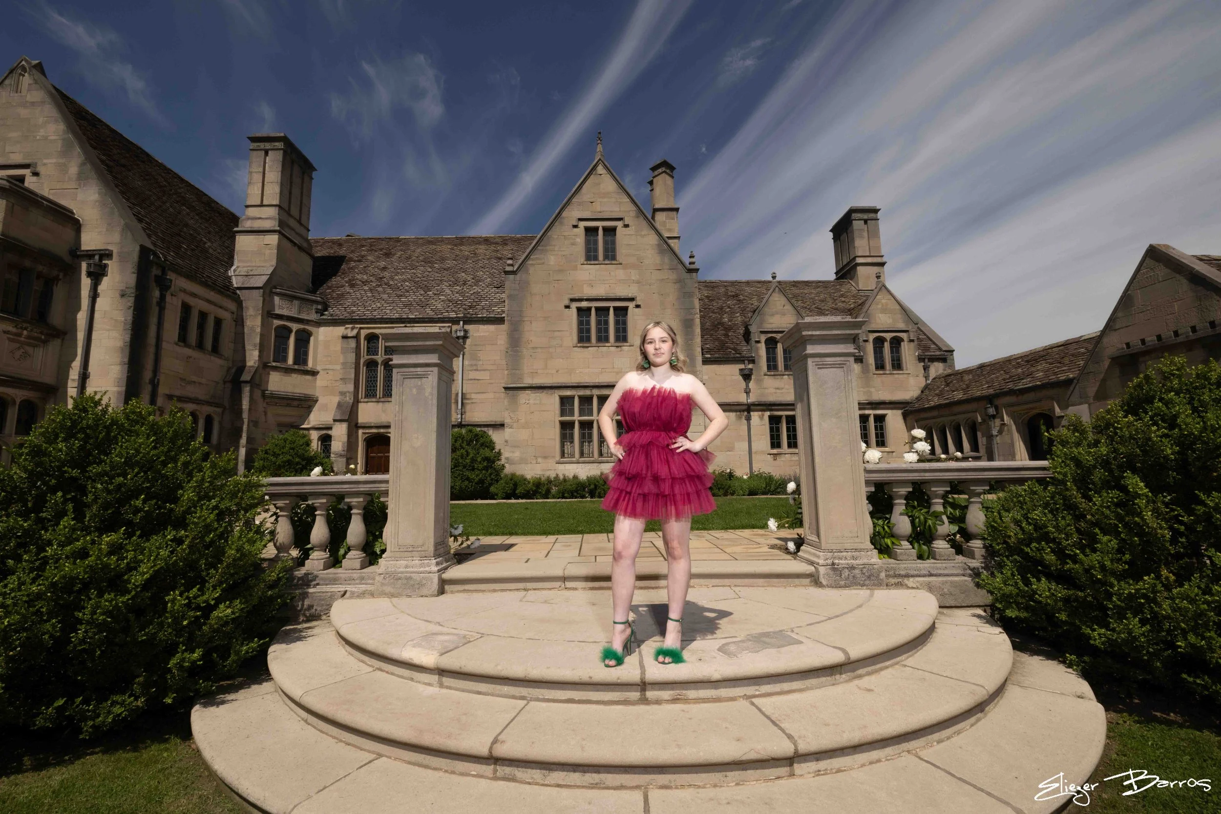 Senior Pictures Pittsburgh at Hartwood Acres Mansion. Young woman in a vibrant pink ruffled dress posing on the grand stone steps and formal gardens in front of the historic mansion for sophisticated outdoor senior pictures.