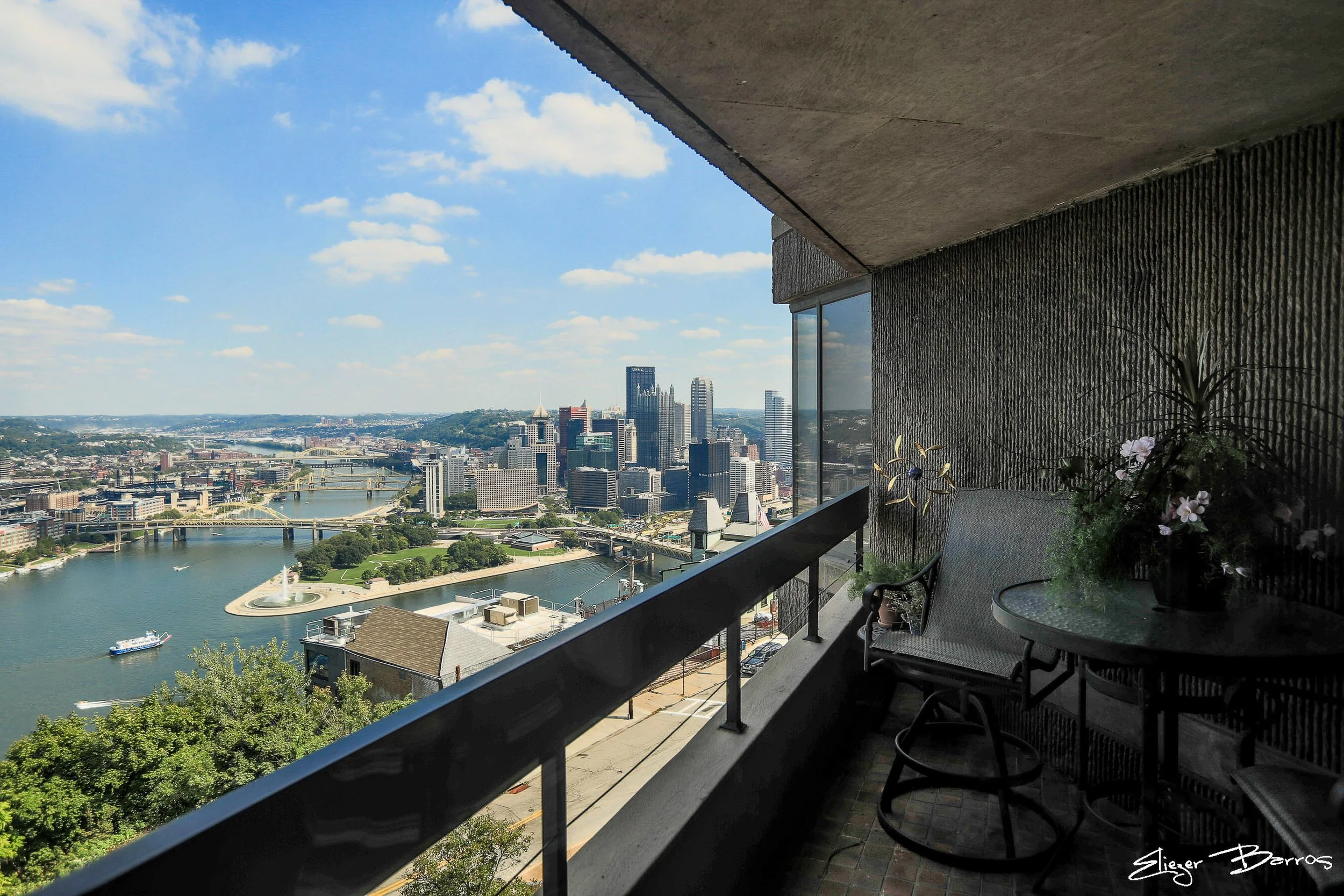 Cityscape view from a balcony showing a river with bridges, tall skyscrapers, and a park under a partly cloudy sky.