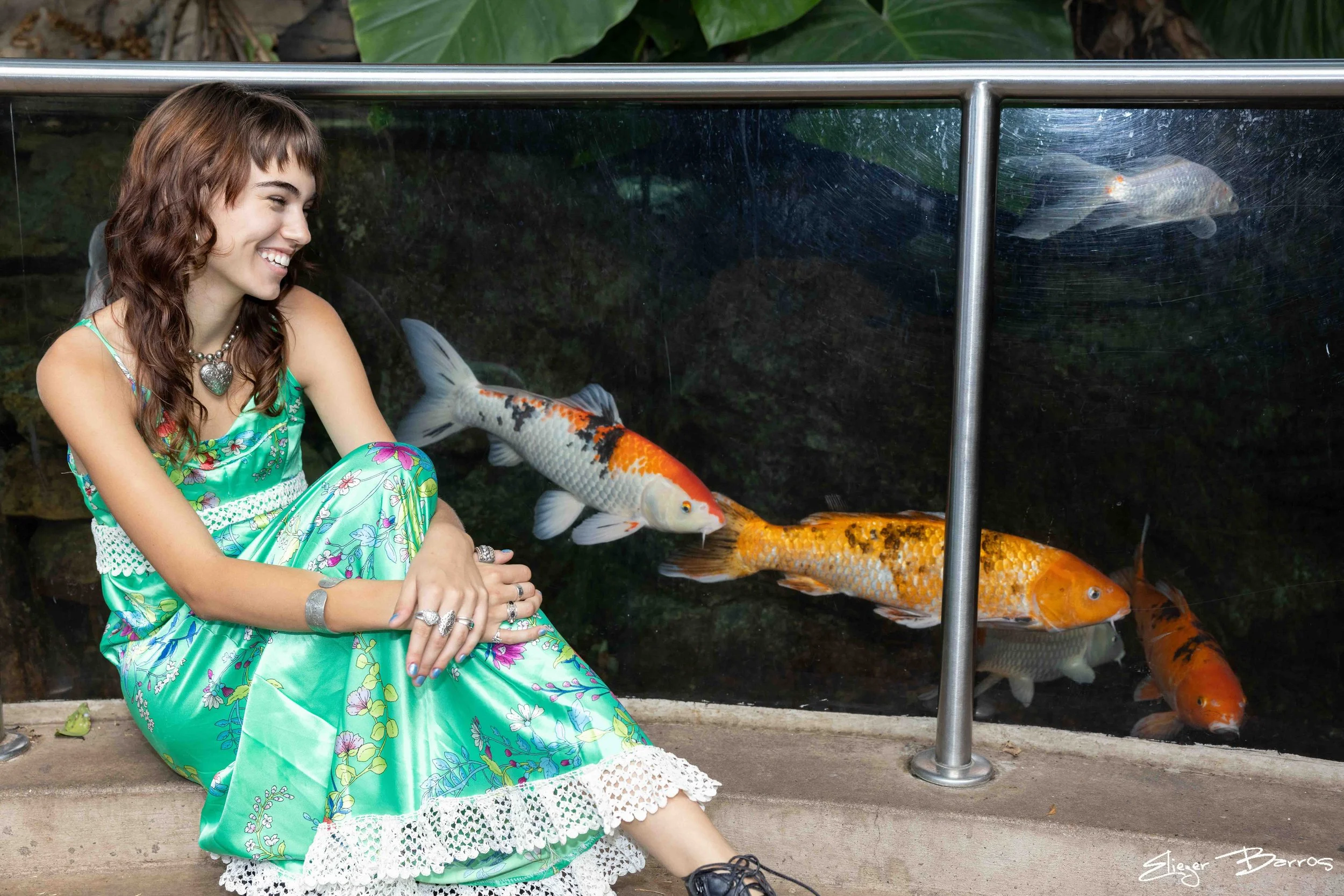 A smiling woman in a green floral dress sitting near a fish tank with goldfish inside.