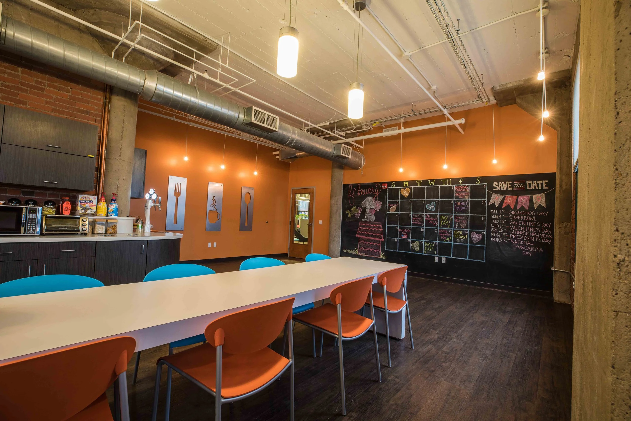 Interior of a modern restaurant or cafe with exposed ductwork, industrial-style lighting, a chalkboard calendar on a black wall, and a kitchen counter with supplies. There are tables and colorful chairs for dining.