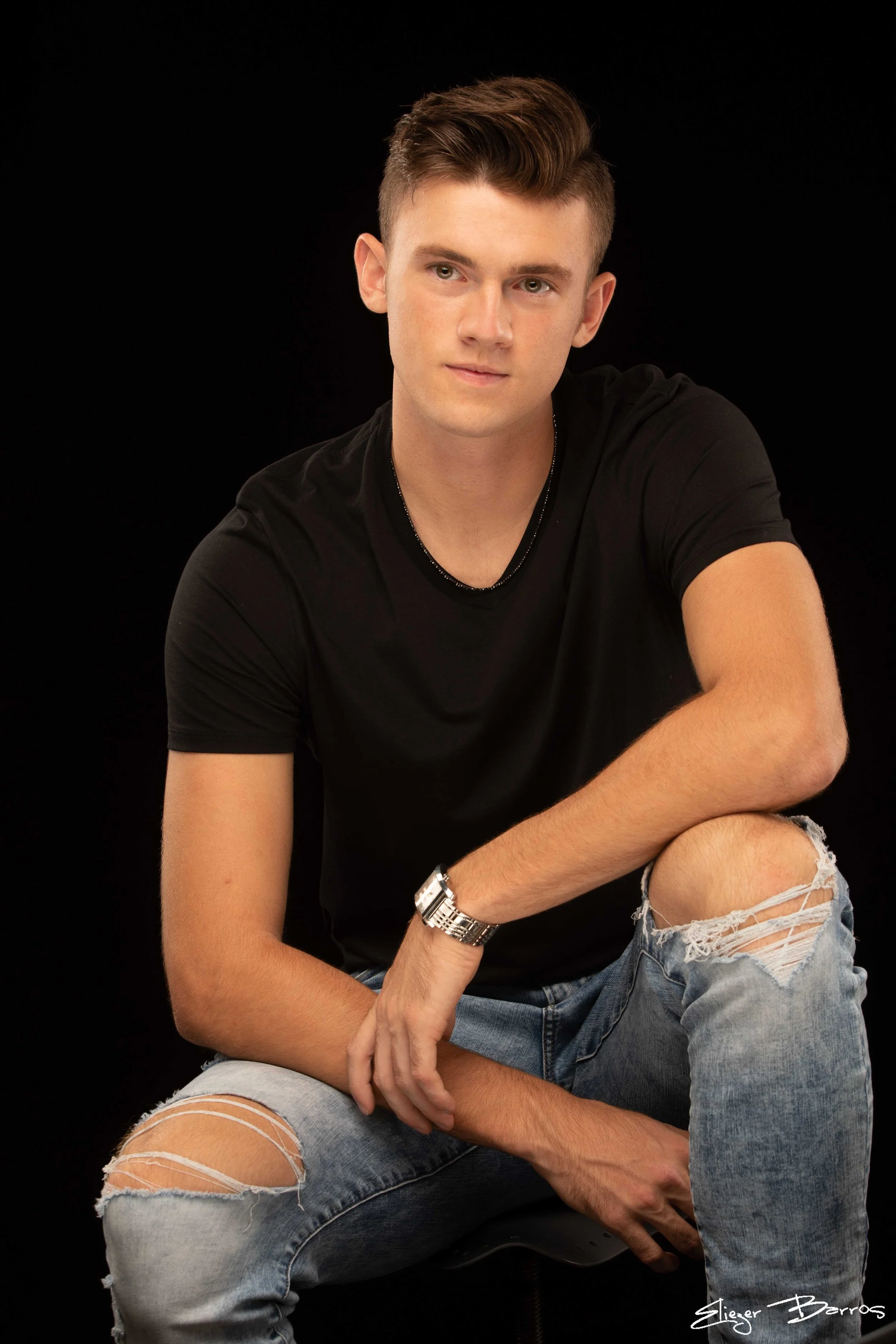 A young man with brown hair and fair skin wearing a black T-shirt and ripped jeans, sitting against a black background, looking towards the camera with a slight smile.