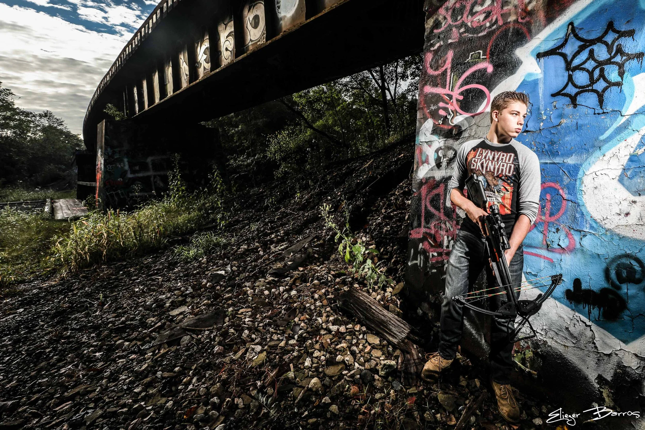 Senior Pictures Pittsburgh at Seldom Seen Greenway. Young man with a crossbow posing under a graffiti-covered bridge in a rugged, wooded setting for unique urban senior pictures.