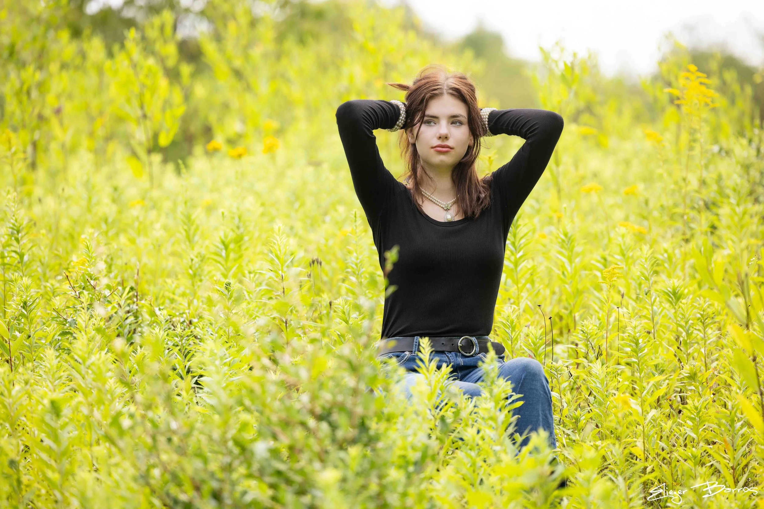 Young woman with brown hair, wearing a black top and jeans, sitting in a green field of tall plants, with her hands resting on her head and looking to the side.