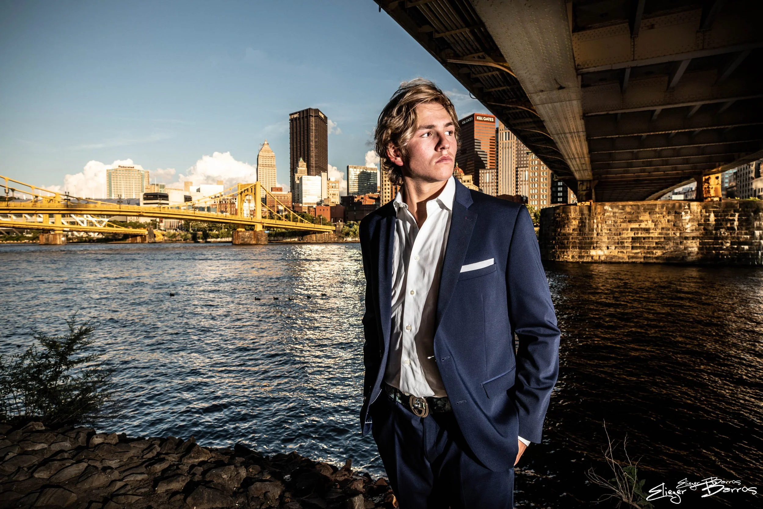 A young man in a navy blue suit and white shirt standing under a bridge by a river, with a city skyline in the background during sunset.