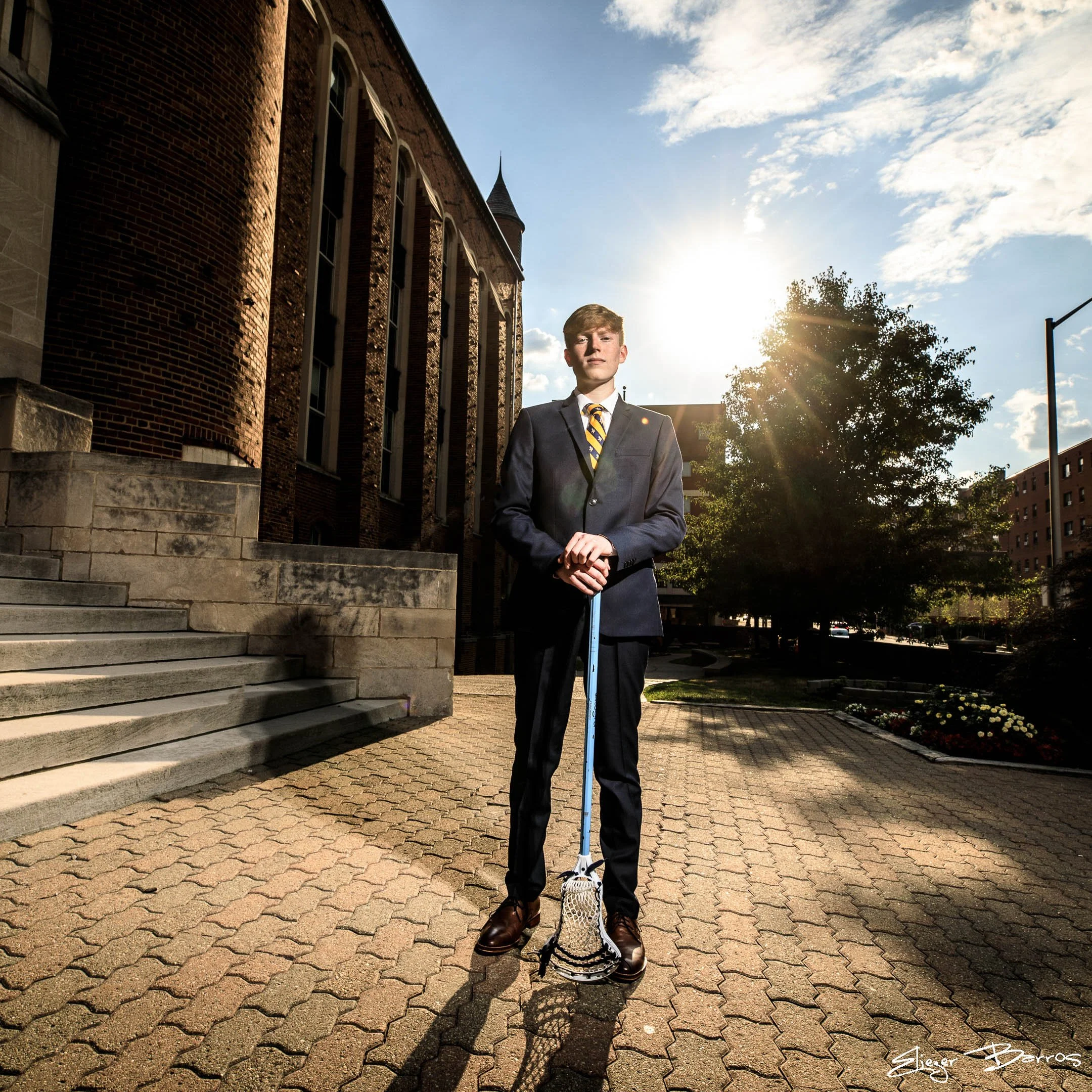 A young man wearing a suit and tie, standing outdoors on a paved area in front of a brick building with steps, holding a scooter. The scene is backlit by the sun, creating a silhouette effect and lens flare. The sky is partly cloudy with a tree and c