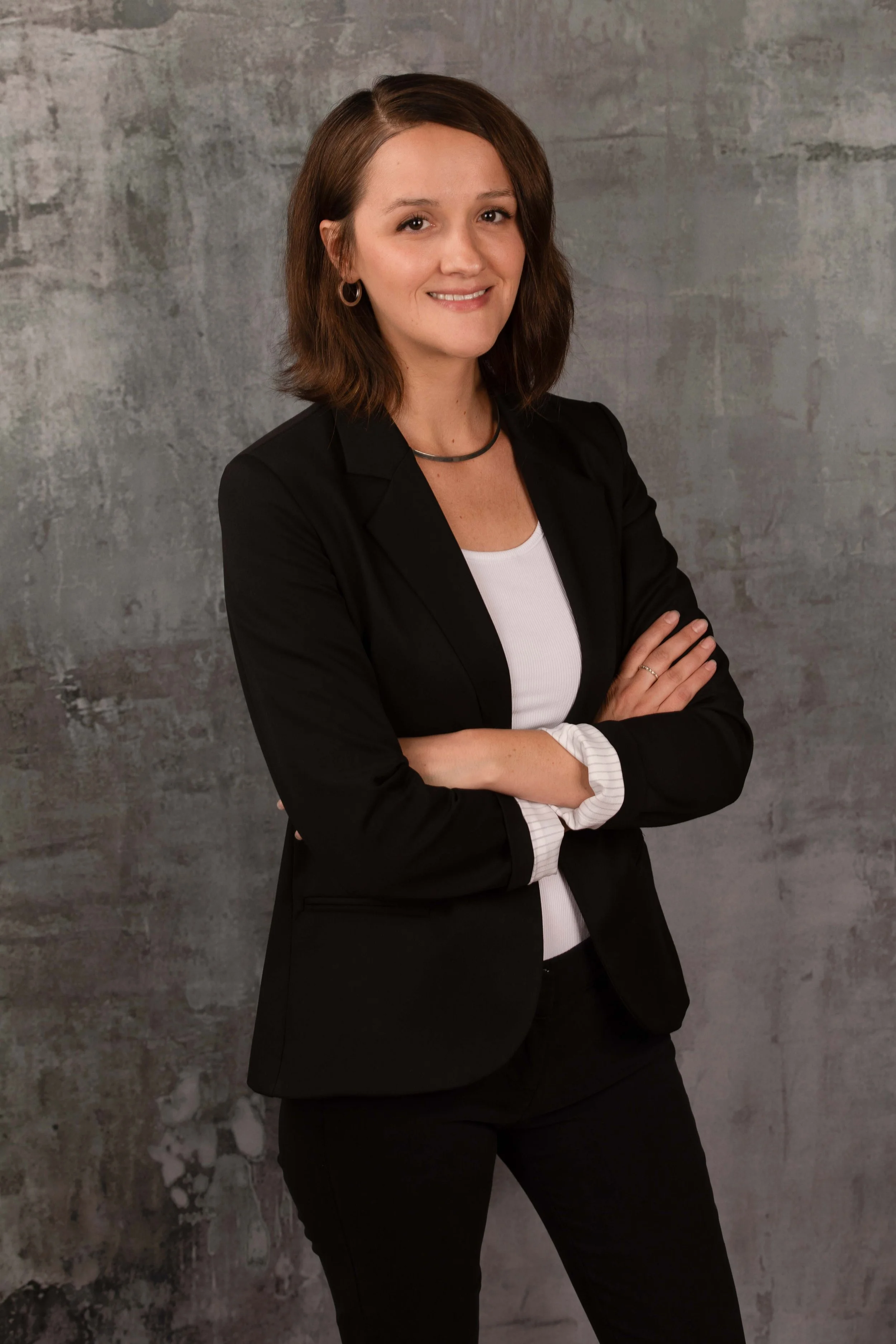 A professional woman with shoulder-length brown hair, wearing a black blazer, white top, and jewelry, standing with arms crossed in front of a concrete-style background.