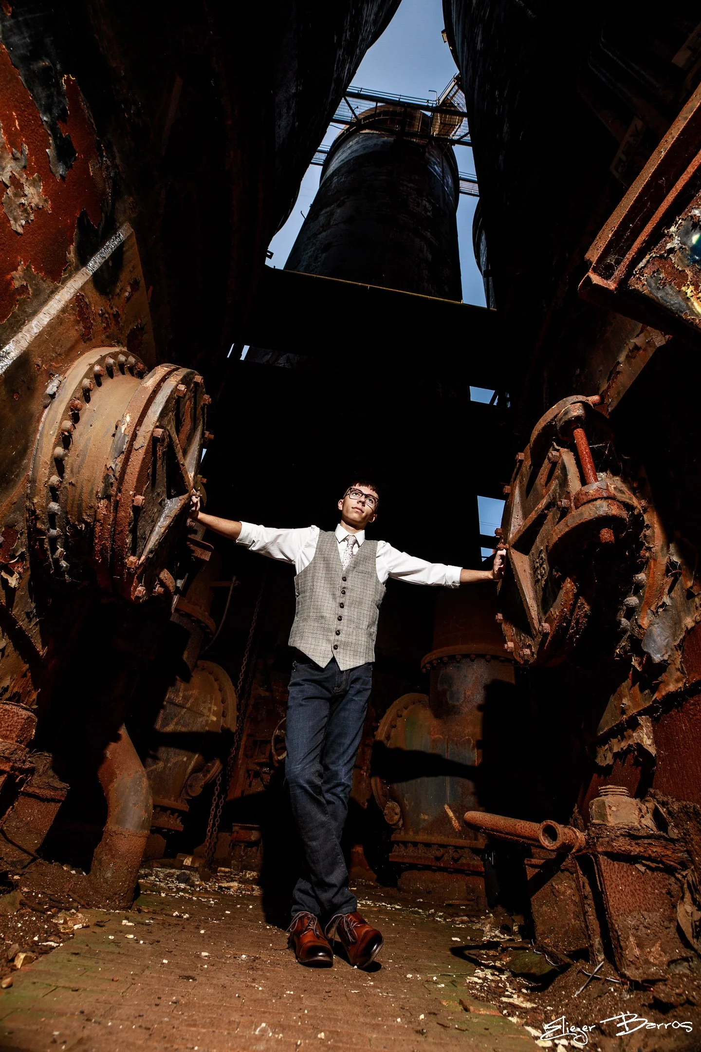 A young man dressed in a vest, white shirt, and dark pants stands with arms outstretched inside a rusty industrial structure, possibly an old factory or shipwreck, with large pipes and machinery around him and a patch of blue sky visible above.