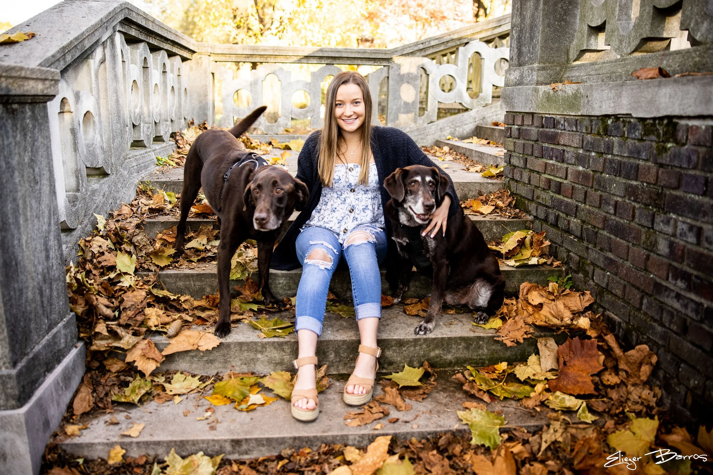 A young woman with long brown hair, wearing a white dress with blue floral pattern, ripped blue jeans, and a black cardigan, sitting on outdoor stone stairs covered with autumn leaves, flanked by two dogs, one brown and one black with white markings,