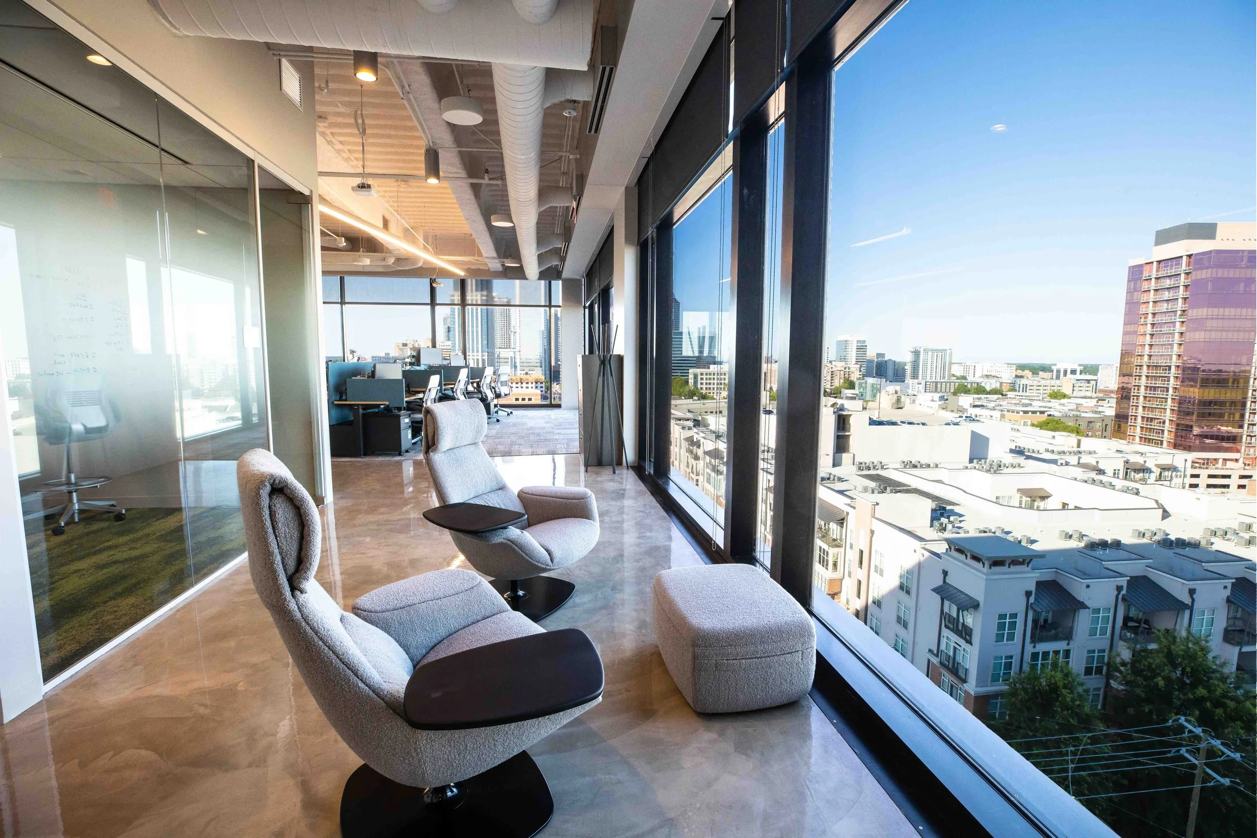 Modern office with large floor-to-ceiling windows showing city buildings and a blue sky, chairs arranged near the windows, and workstations in the background.