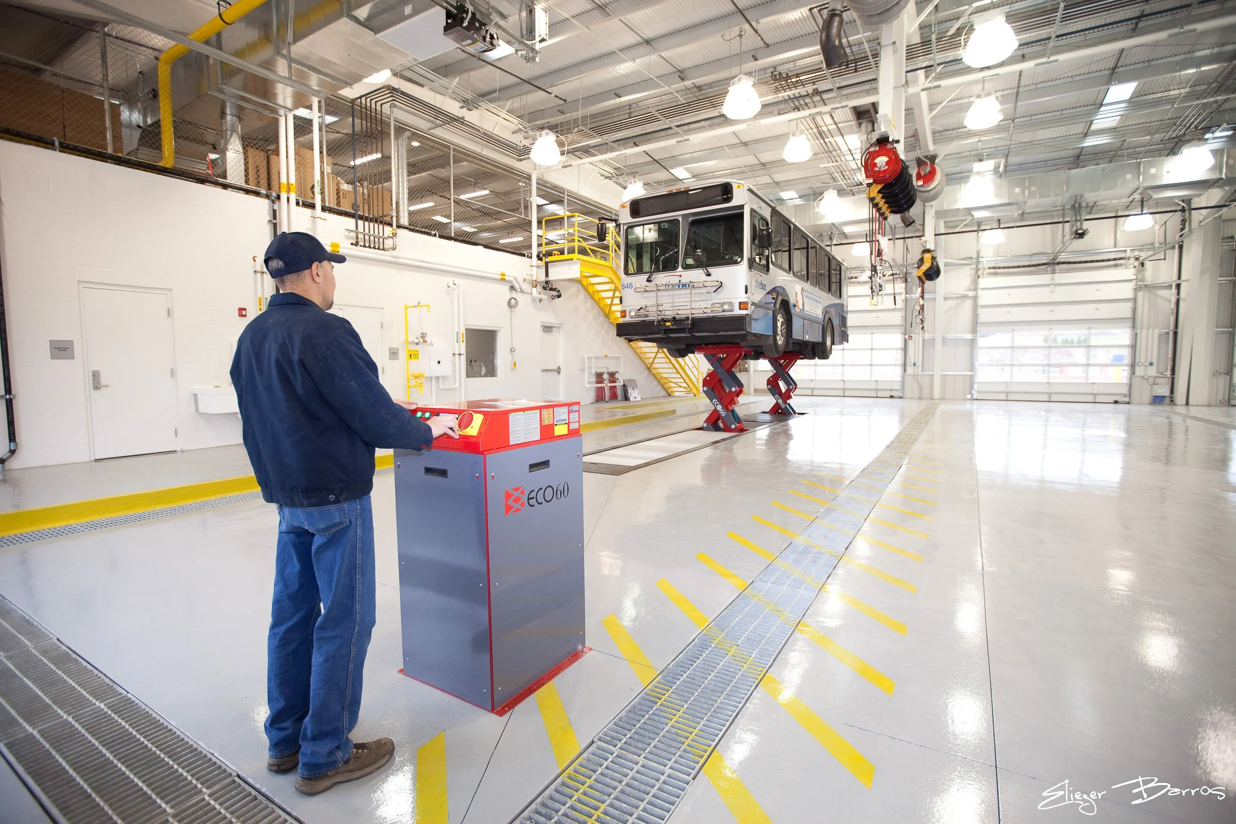 A worker in a blue jacket and cap operates a control panel next to a bus lifted on hydraulic lifts inside a spacious, well-lit industrial workshop with high ceilings, yellow safety markings on the floor, and storage shelves overhead.