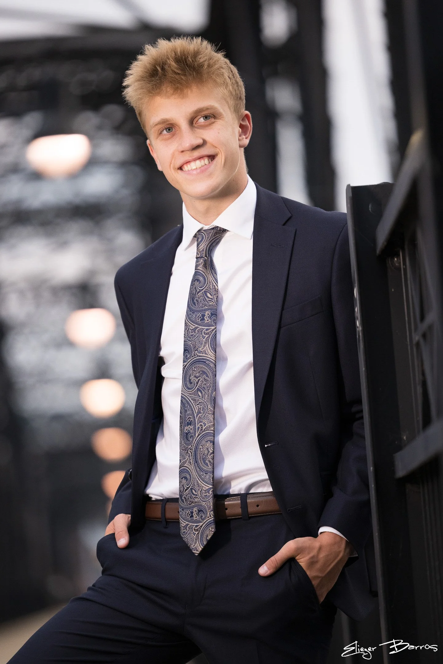 A young man in a dark suit and patterned tie, smiling confidently, standing with his hand on his hip in an urban setting.