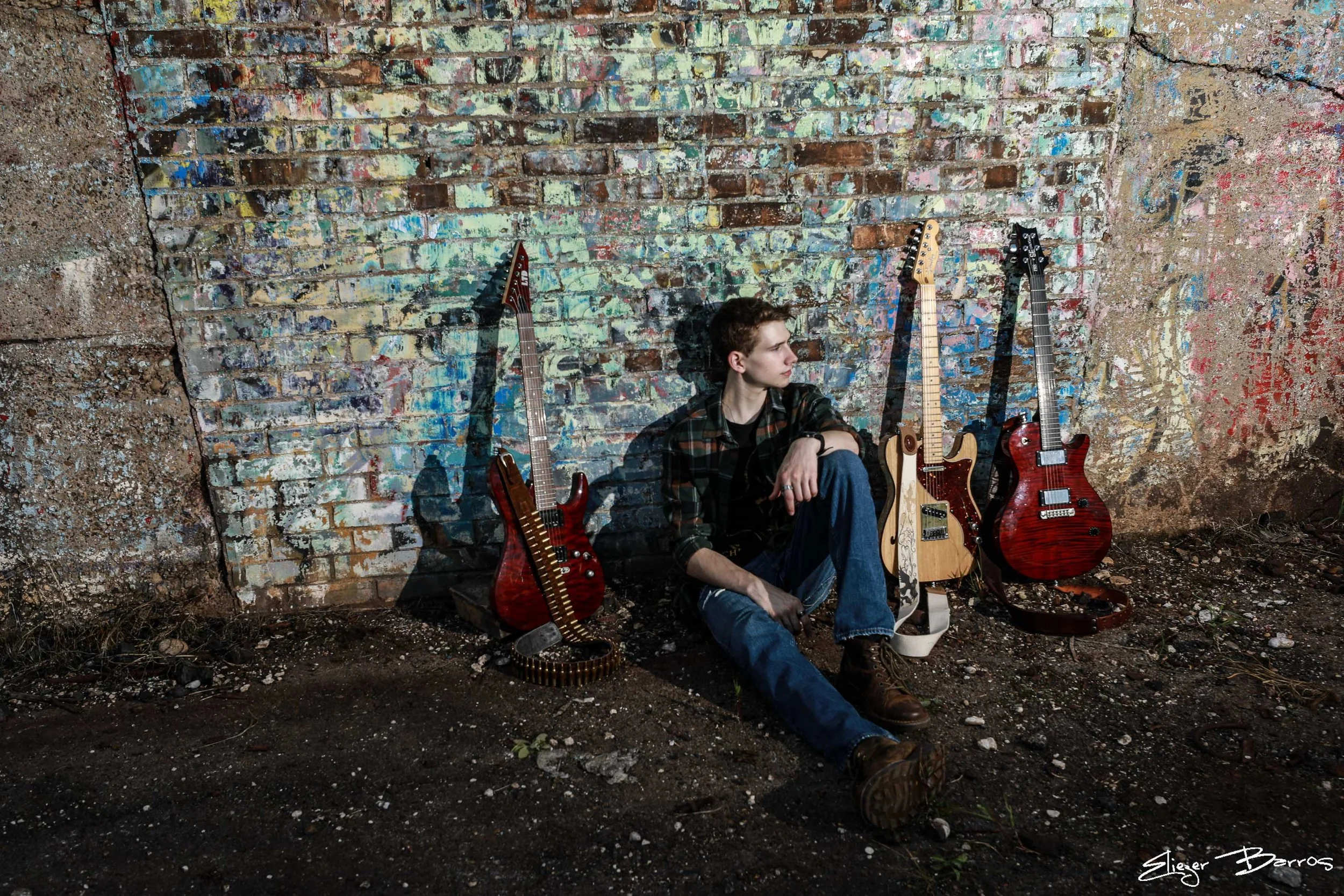 Young man sitting on the ground against a colorful, graffiti-covered brick wall with four electric guitars resting nearby.