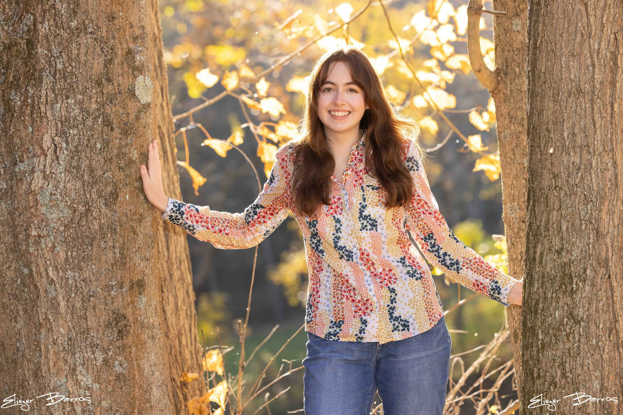 A young woman with long brown hair smiling and standing between two large trees in autumn with yellow leaves.