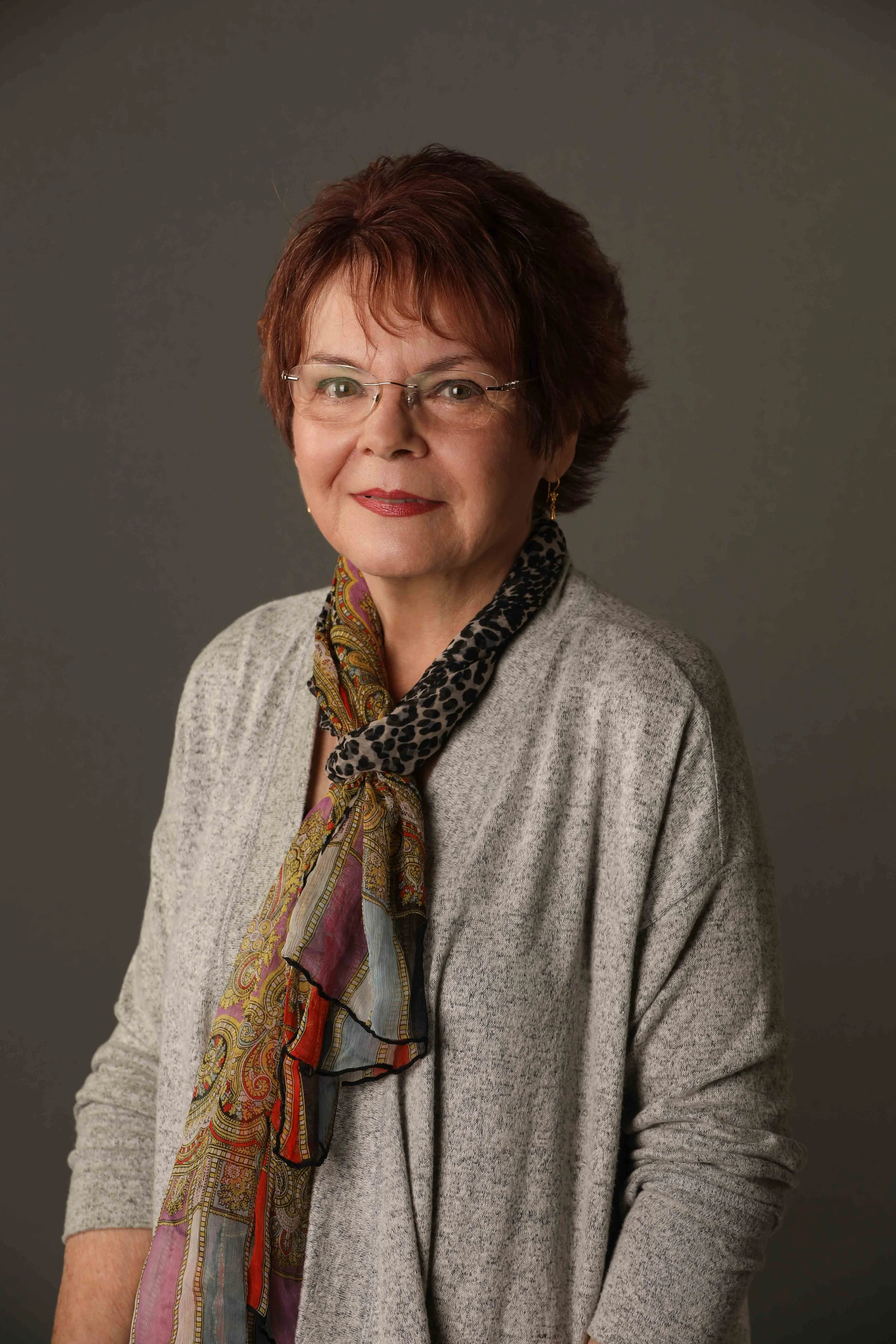 A portrait of an older woman with short, reddish-brown hair, wearing glasses, a light gray sweater, and a colorful scarf around her neck, against a dark gray background.
