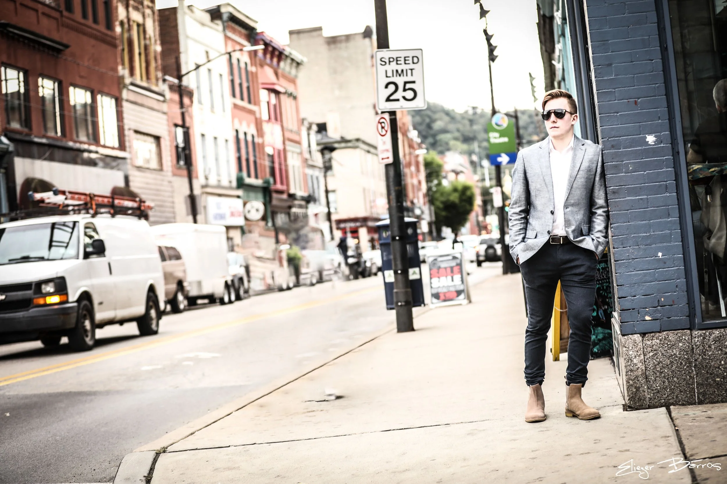 A young man with sunglasses in a gray blazer and black pants is standing on a city sidewalk, leaning against a dark gray brick building. The background shows parked cars, a street with a speed limit sign of 25 miles per hour, and colorful buildings w
