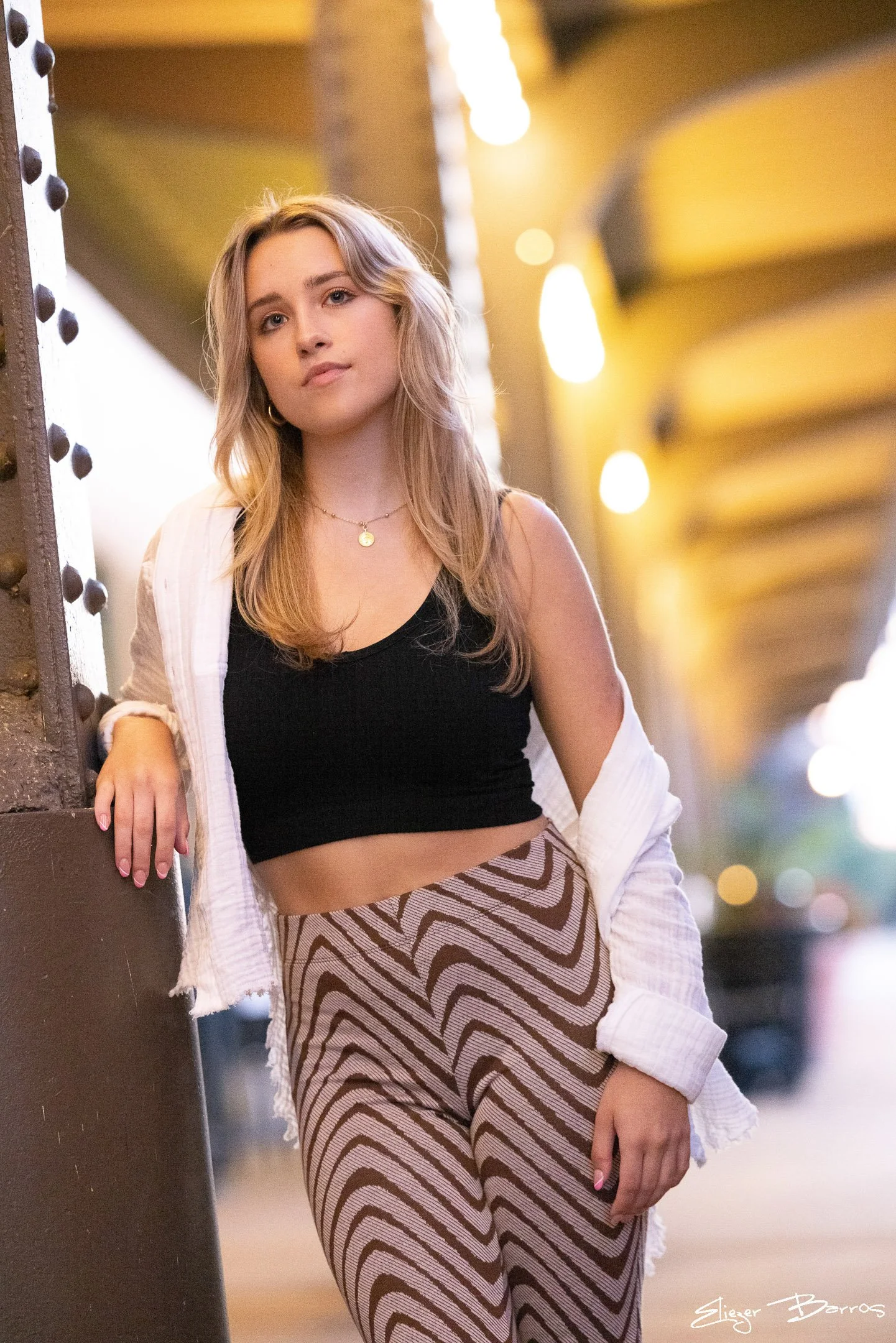 A young woman with blonde hair, wearing a black crop top, earrings, a necklace, and patterned pants, leaning against a wall in a well-lit indoor setting.