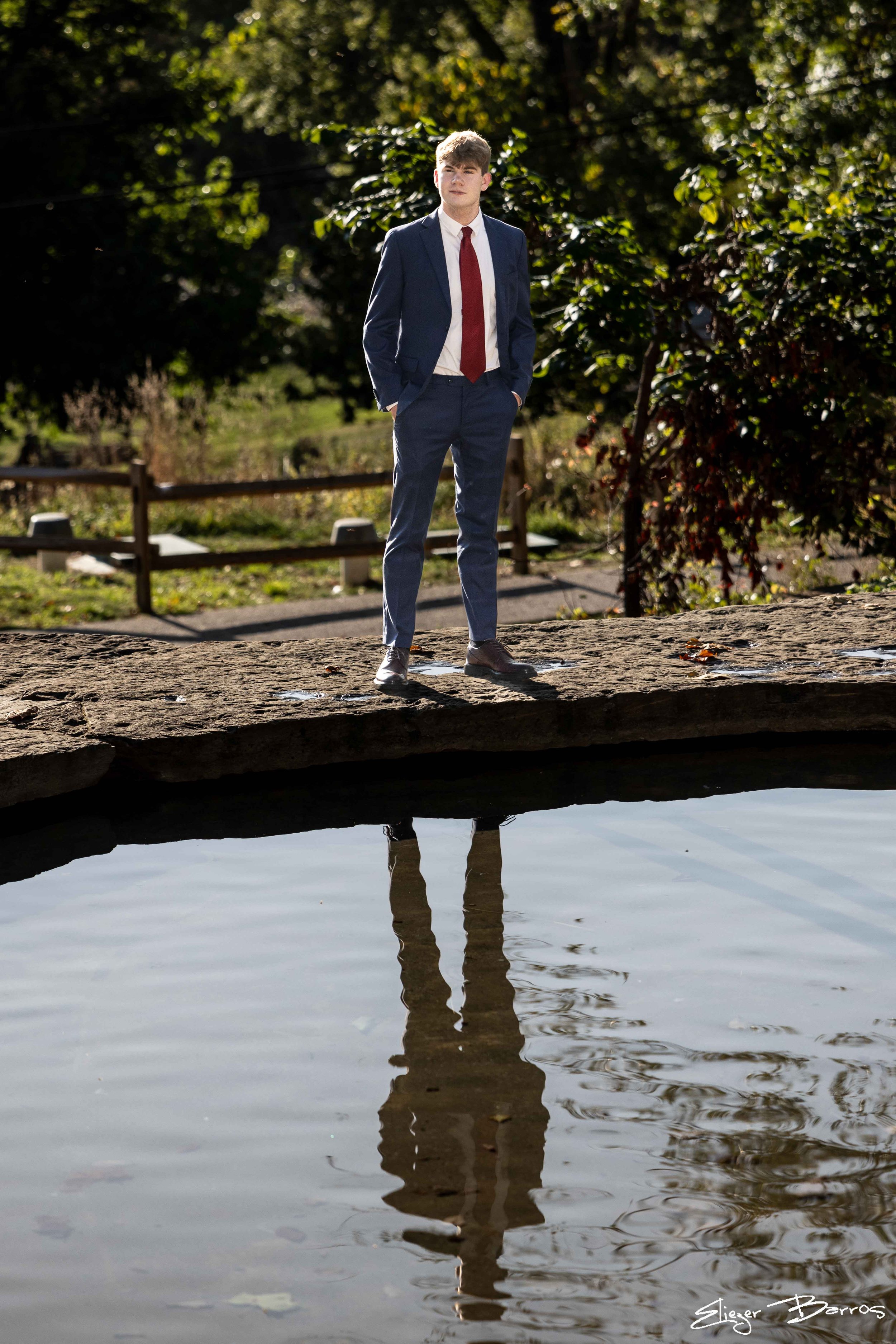 A young man in a navy suit with a white shirt and red tie stands outdoors on a rock near a calm body of water, with trees and sunlight in the background. His reflection is visible in the water.