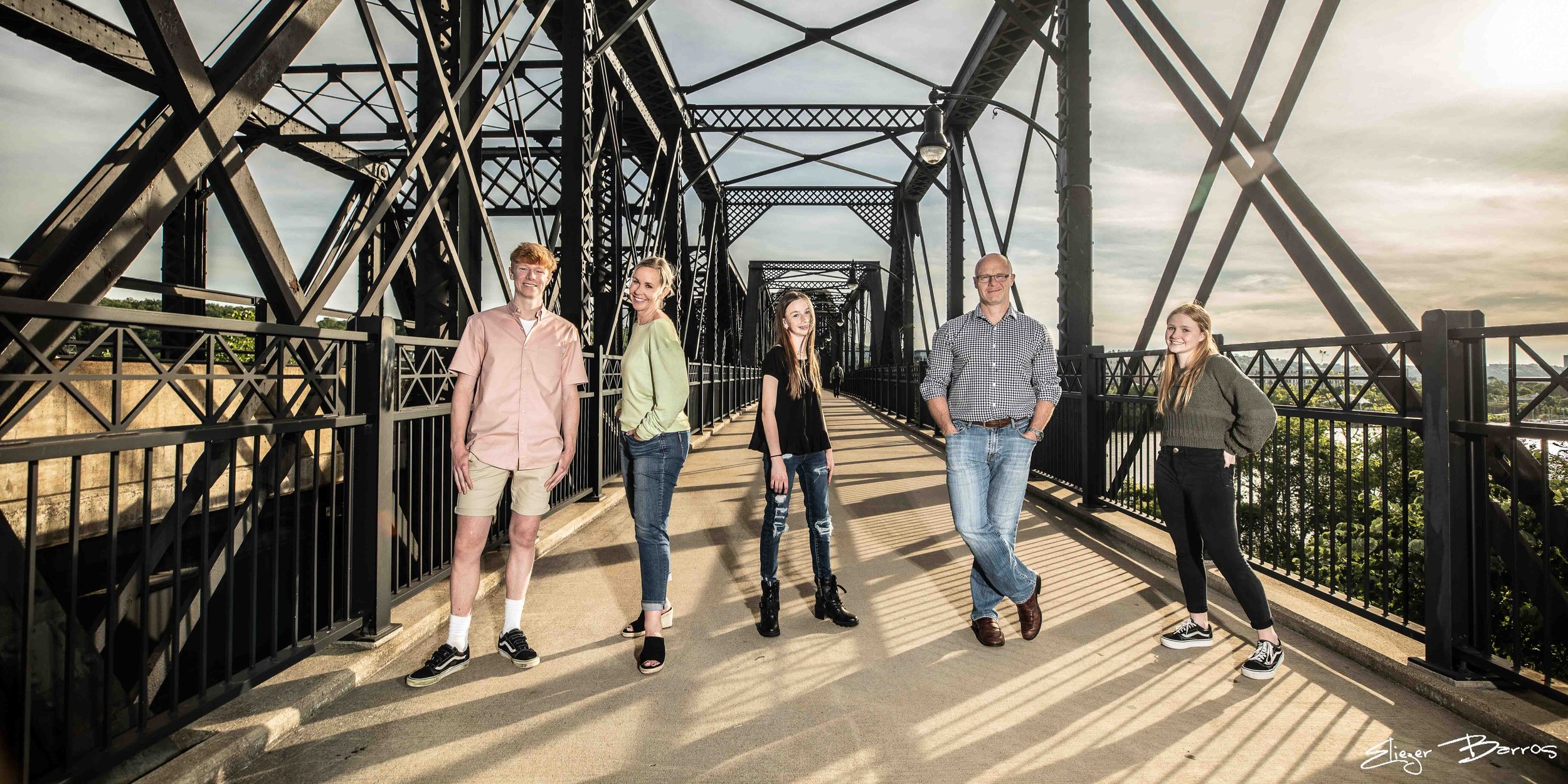 A group of five young people standing on a bridge with steel framework, smiling and posing for a photo during sunset.