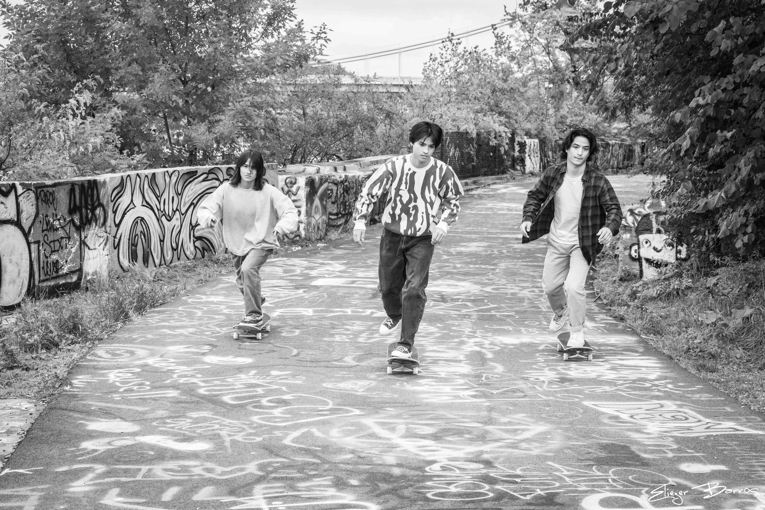 Three young people skateboarding on a graffiti-covered pathway with trees on either side in black and white.