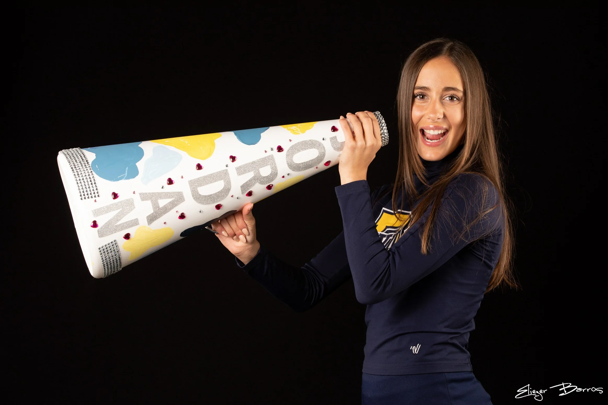 A young woman with long brown hair holding a large celebratory party cone decorated with colorful hearts and patterns, smiling and standing against a black background.
