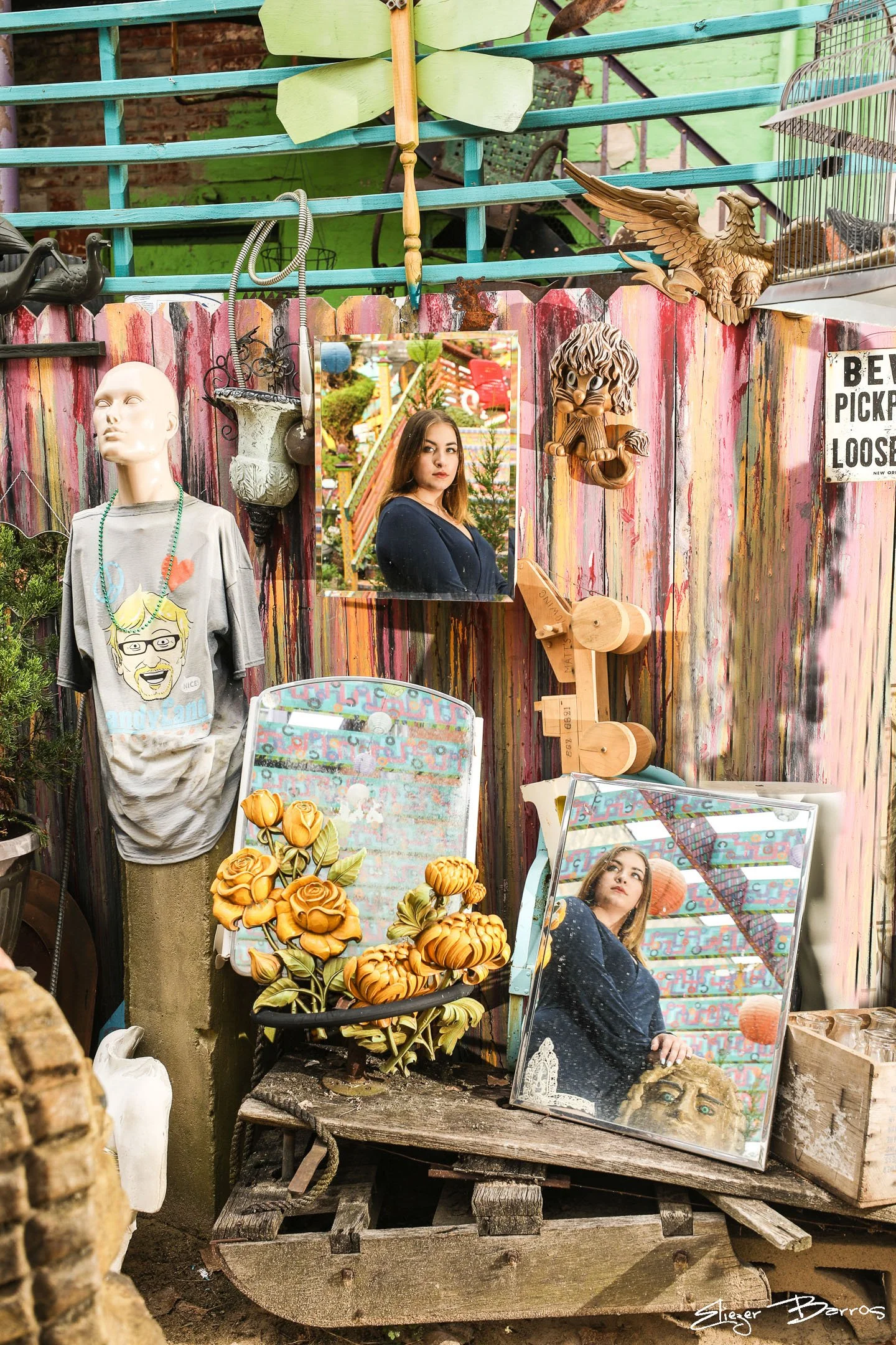 Decorative outdoor scene with a mannequin head wearing a graphic T-shirt, two mirrors showing a woman with brown hair, and various ornaments including a carved eagle, a lion sculpture, vintage signs, and artificial flowers, all set against a painted 