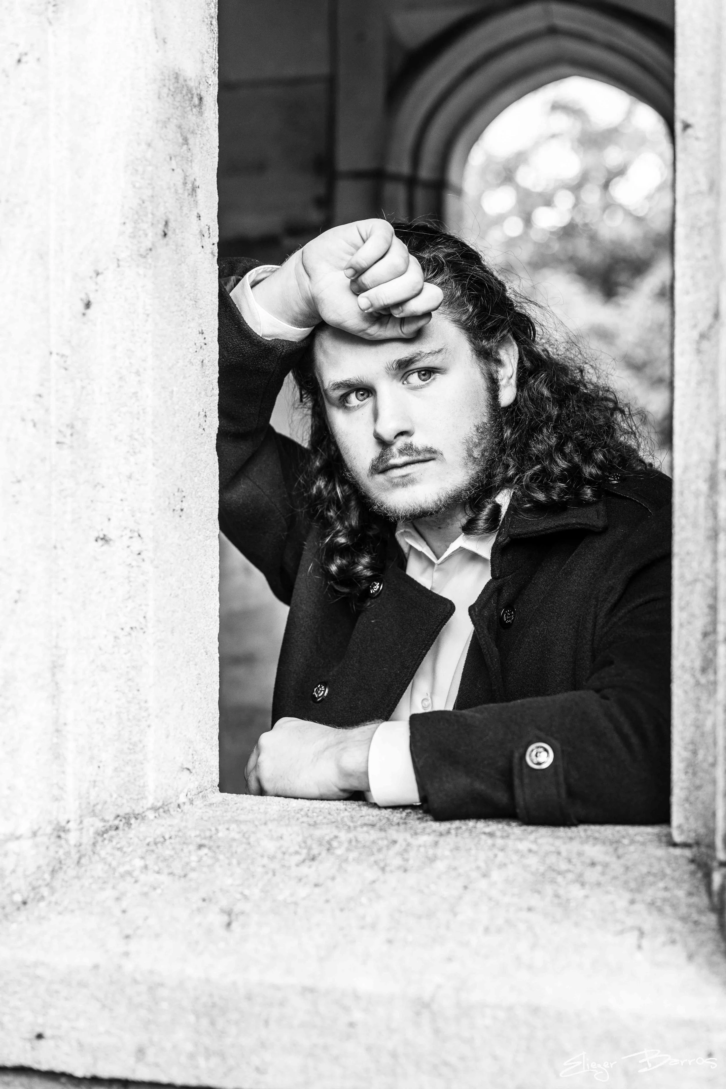 Black and white portrait of a young man with long, curly hair, leaning on a stone window ledge with his hand resting on his forehead. He is wearing a dark coat and a white shirt, looking contemplative.