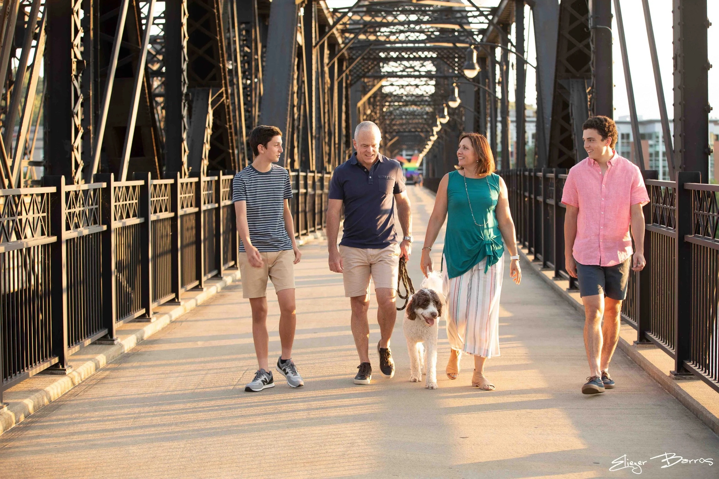 A group of five people walking on a bridge with a dog during sunset. The group includes a man, a woman, and three young men, all smiling and talking.
