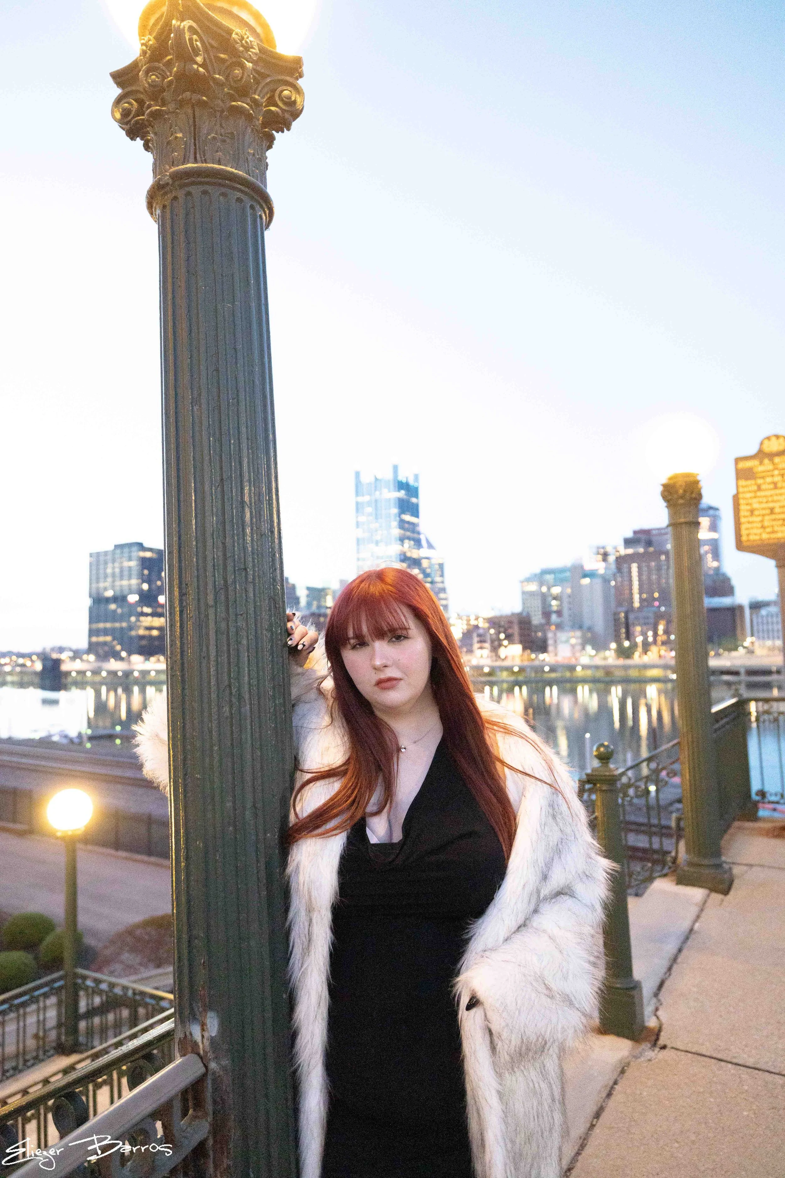 A woman with long red hair and bangs posing on a bridge at dusk, with city buildings and a river in the background. She is wearing a black top and a white fur coat, leaning against a tall street lamp.