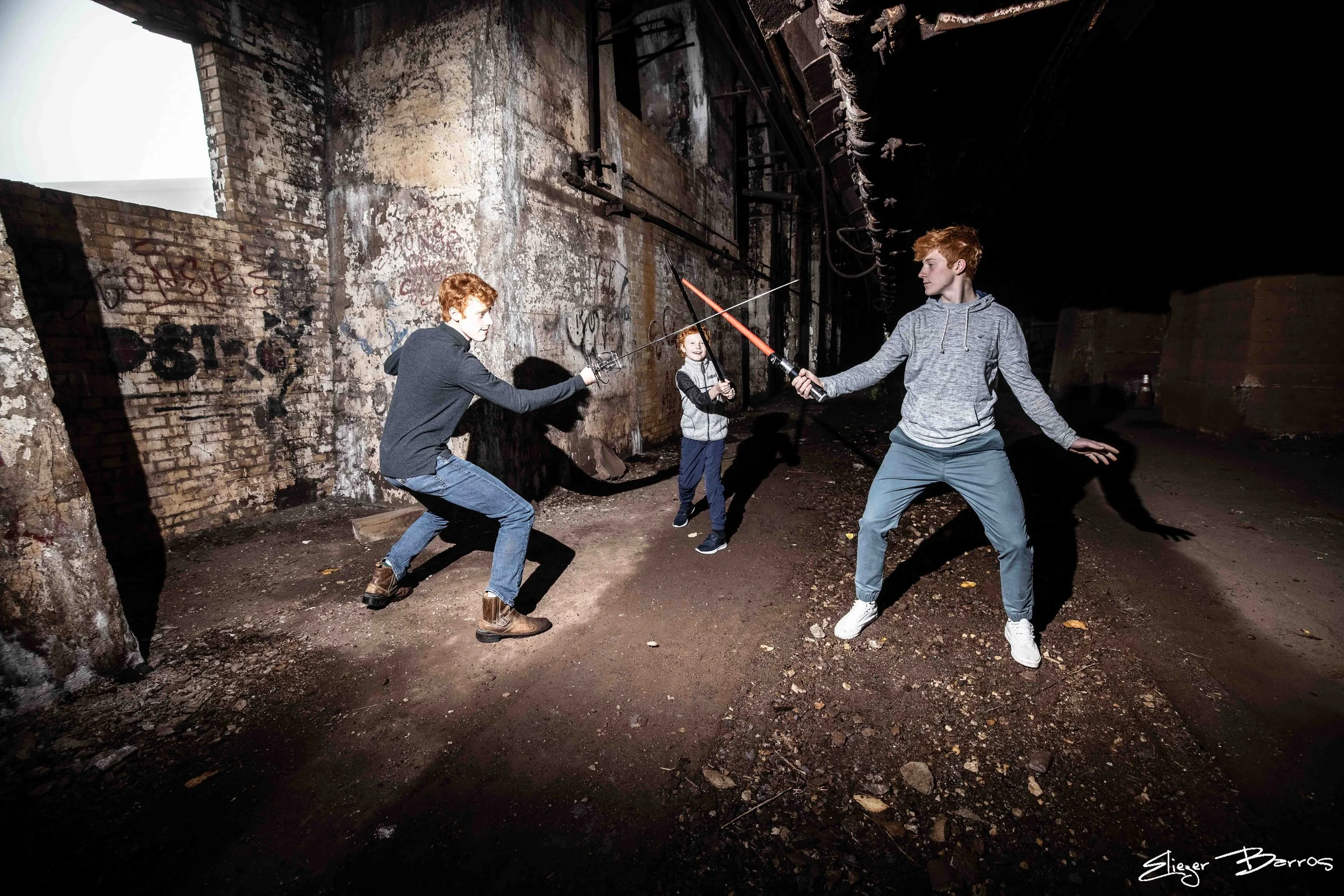 Three young men with red hair fencing in an abandoned, graffiti-covered industrial space with dark, damaged brick walls.