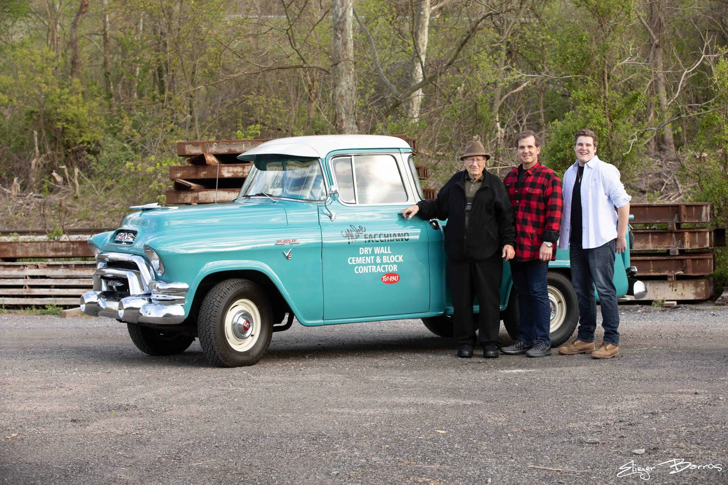 Three men stand next to a vintage blue GMC truck with services written on the door, set against a background of trees and stacked rusty metal beams.
