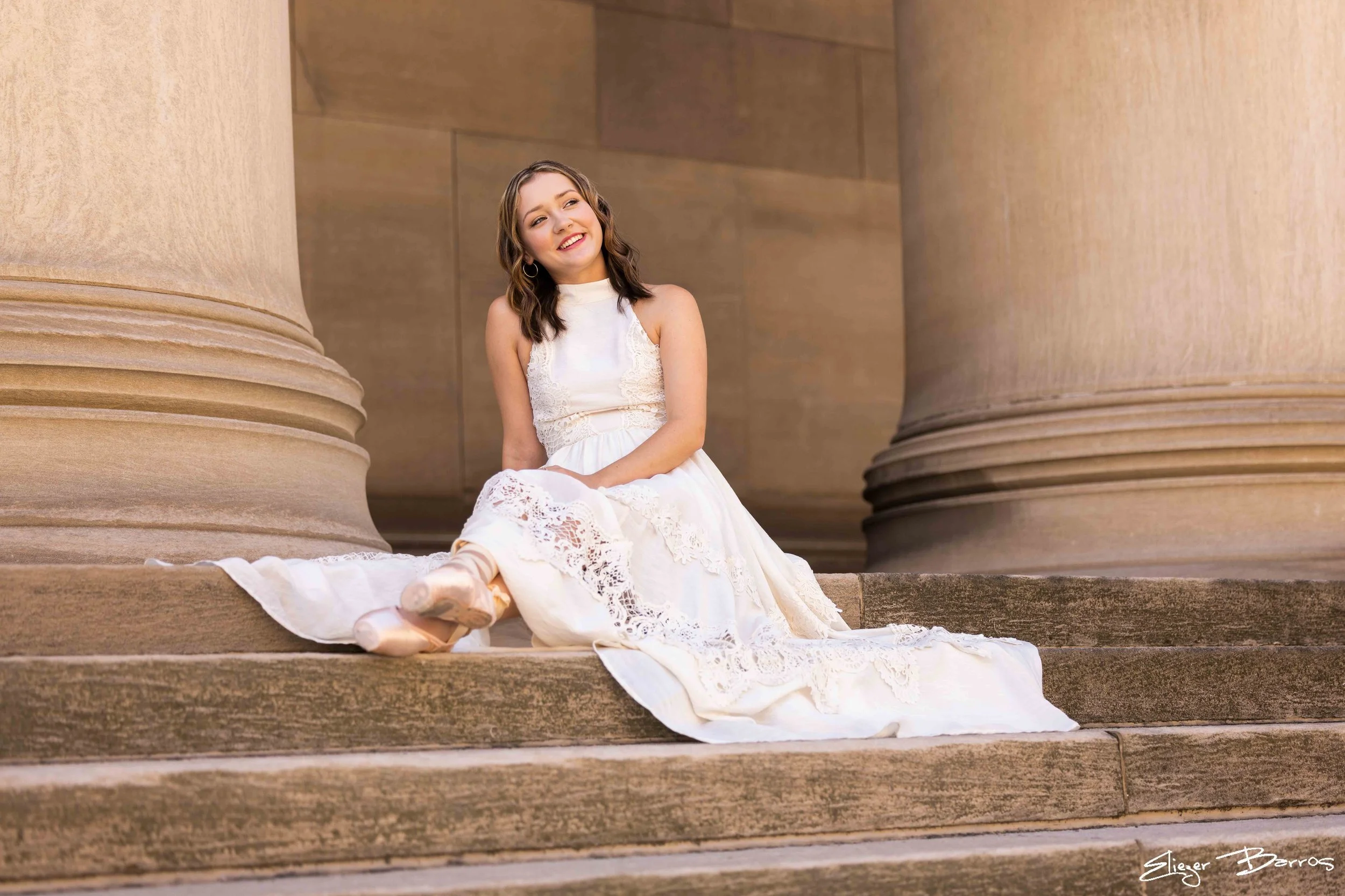 A young woman sitting on steps between large stone columns, wearing a white lace dress and ballet slippers, smiling and looking away.