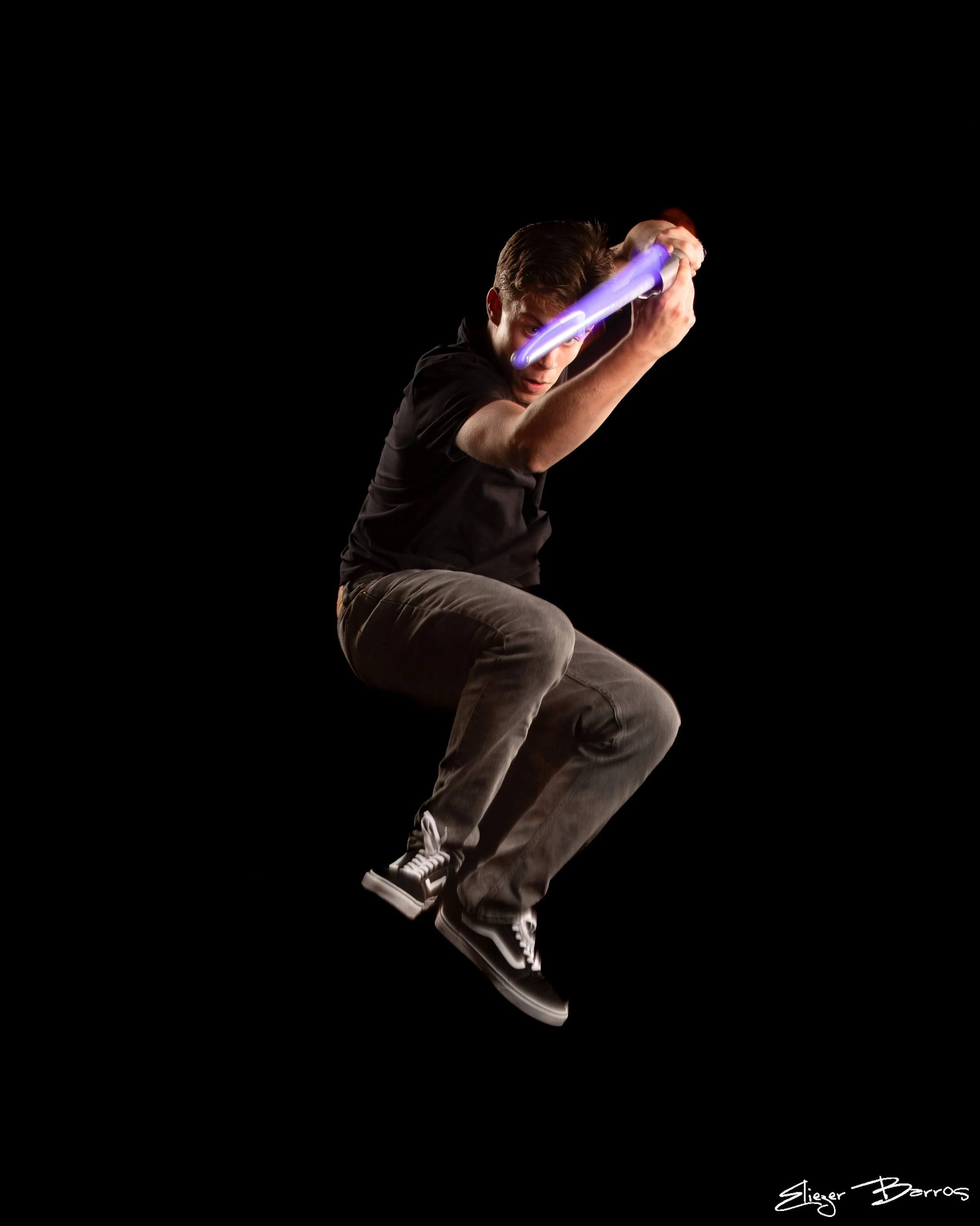 A young man with brown hair dressed in a black shirt and gray pants is holding a glowing purple lightsaber with both hands, posing as if ready for combat, against a plain black background.