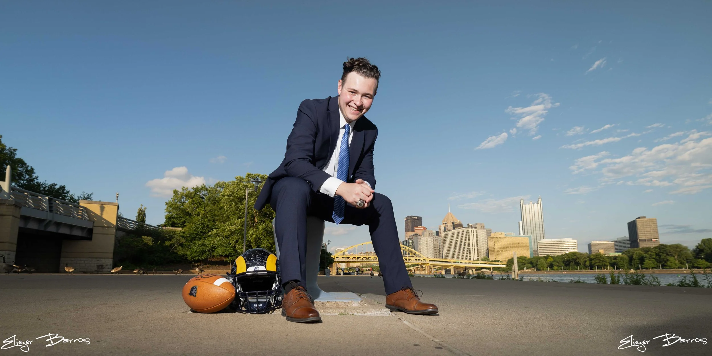 A man in a navy suit crouching on a city sidewalk, smiling, with football helmets and a football on the ground next to him, with a city skyline, including tall buildings, a bridge, and trees, in the background on a clear day.