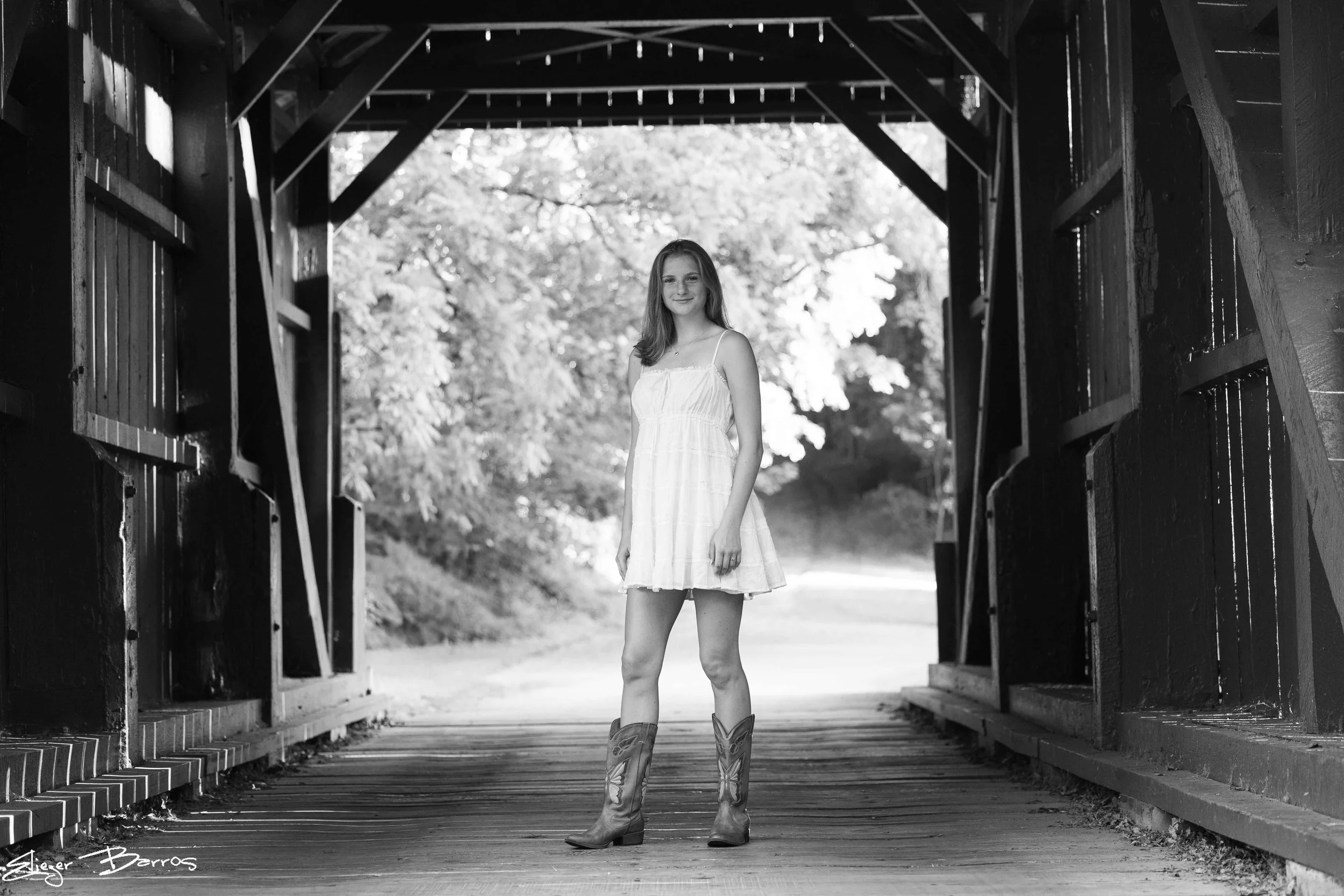 A young woman in a white dress and cowboy boots stands under a wooden covered bridge with a scenic background of trees.