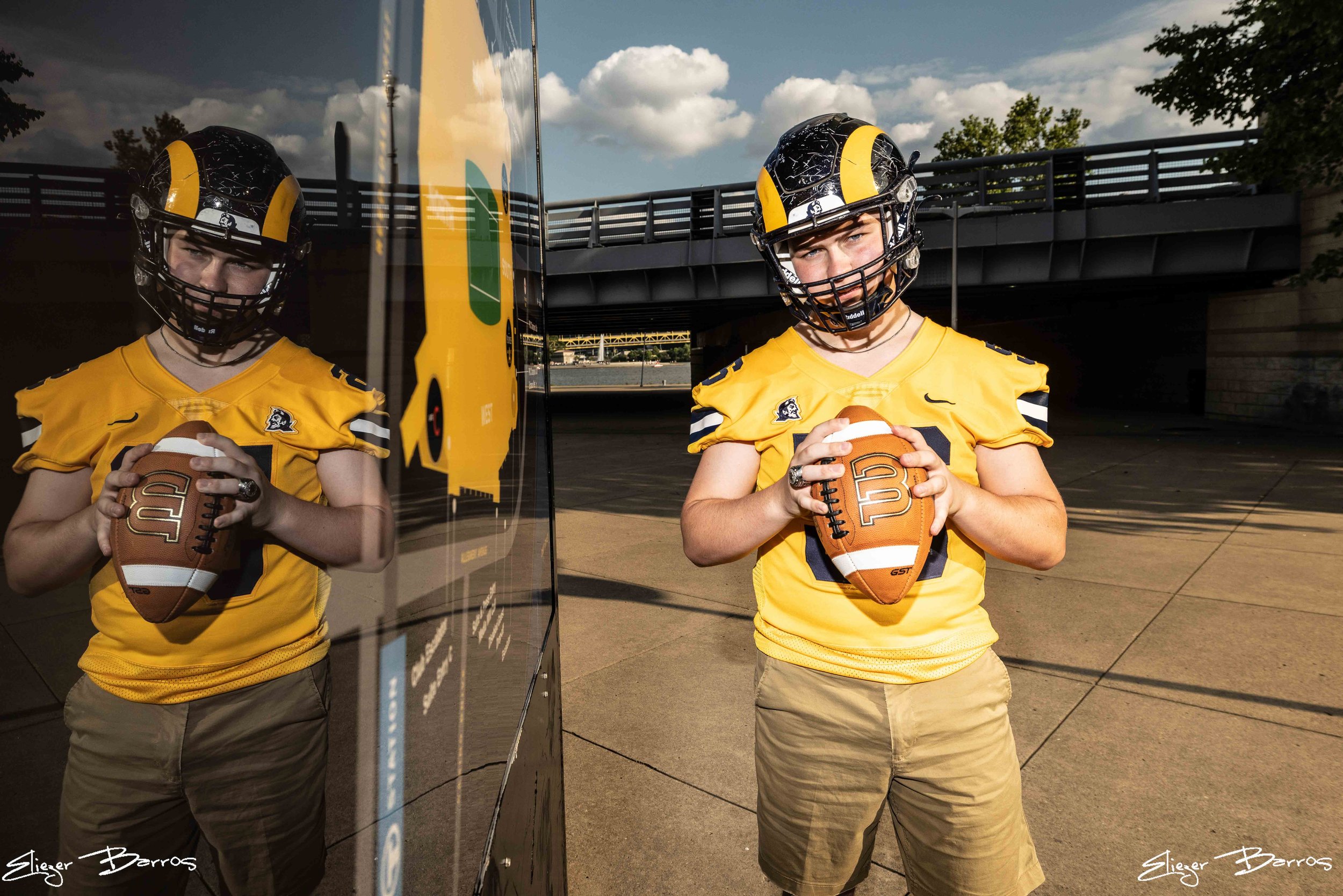 A young man wearing a yellow football uniform and helmet holding a football, standing outdoors near a reflective glass surface with a city bridge and cloudy sky in the background.