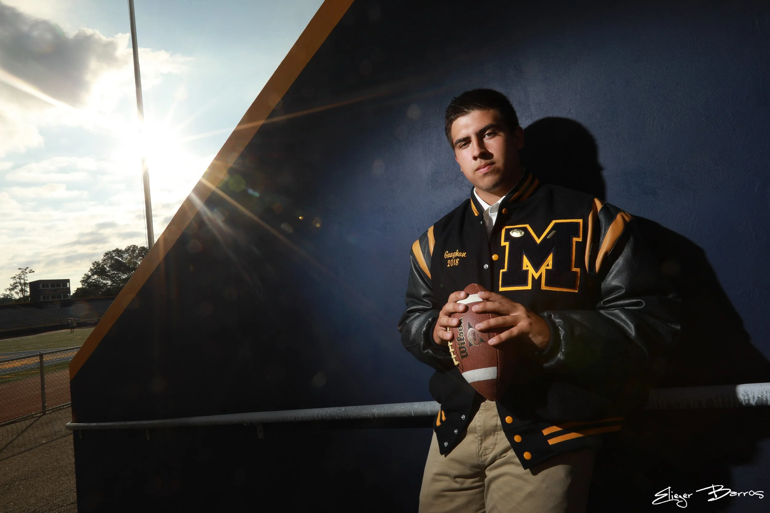 Young man wearing a black and gold jacket with varsity-style lettering, holding a football, standing outdoors with a stadium and cloudy sky in the background, sunlight creating a lens flare.