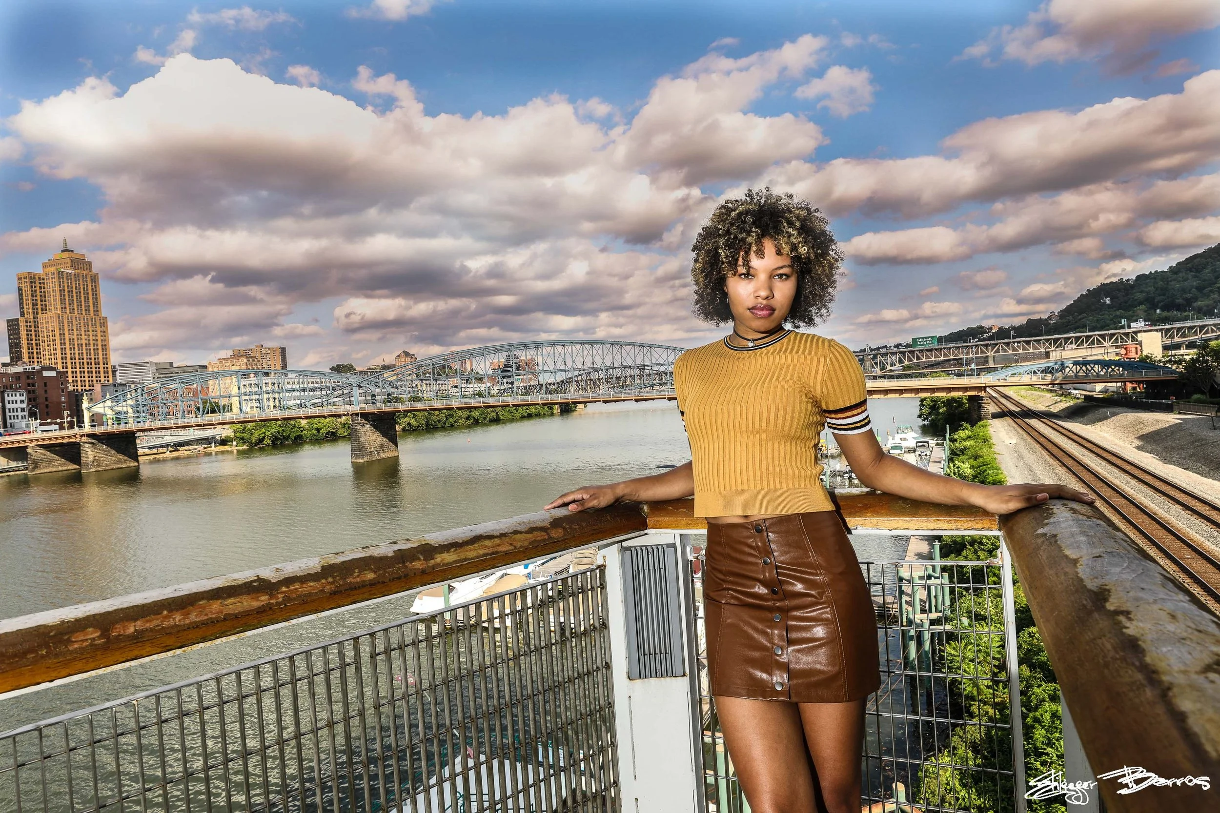 A woman with curly hair wearing a yellow ribbed top and brown leather skirt, standing on a bridge over a river in a city, with bridges, buildings, and a hill in the background under a partly cloudy sky.