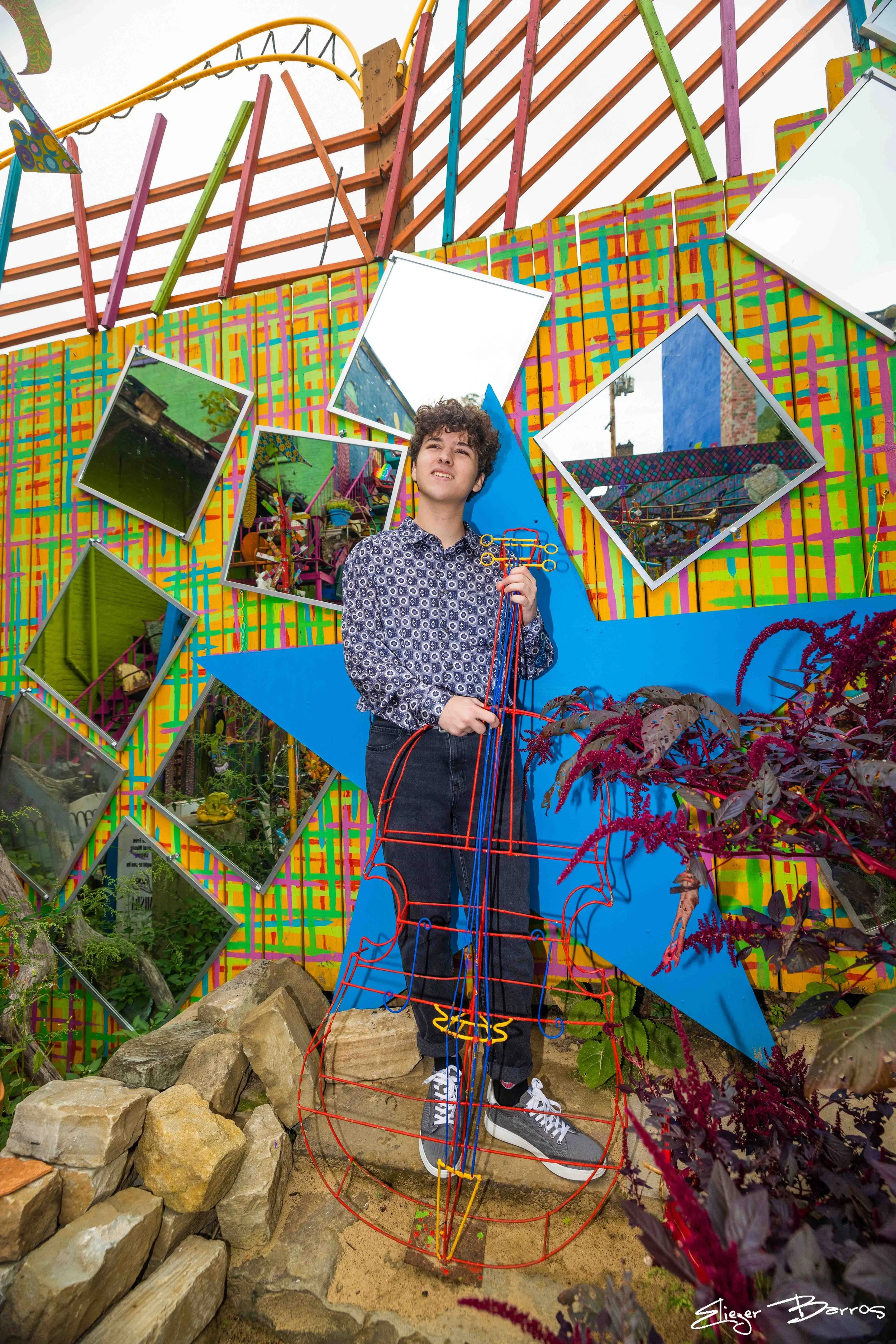 A young man with curly hair and a patterned shirt stands in front of a colorful, artistic backdrop featuring geometric mirrors, vibrant paint patterns, and amusement park rides in the background. He holds a small object in his hand and standing on a 