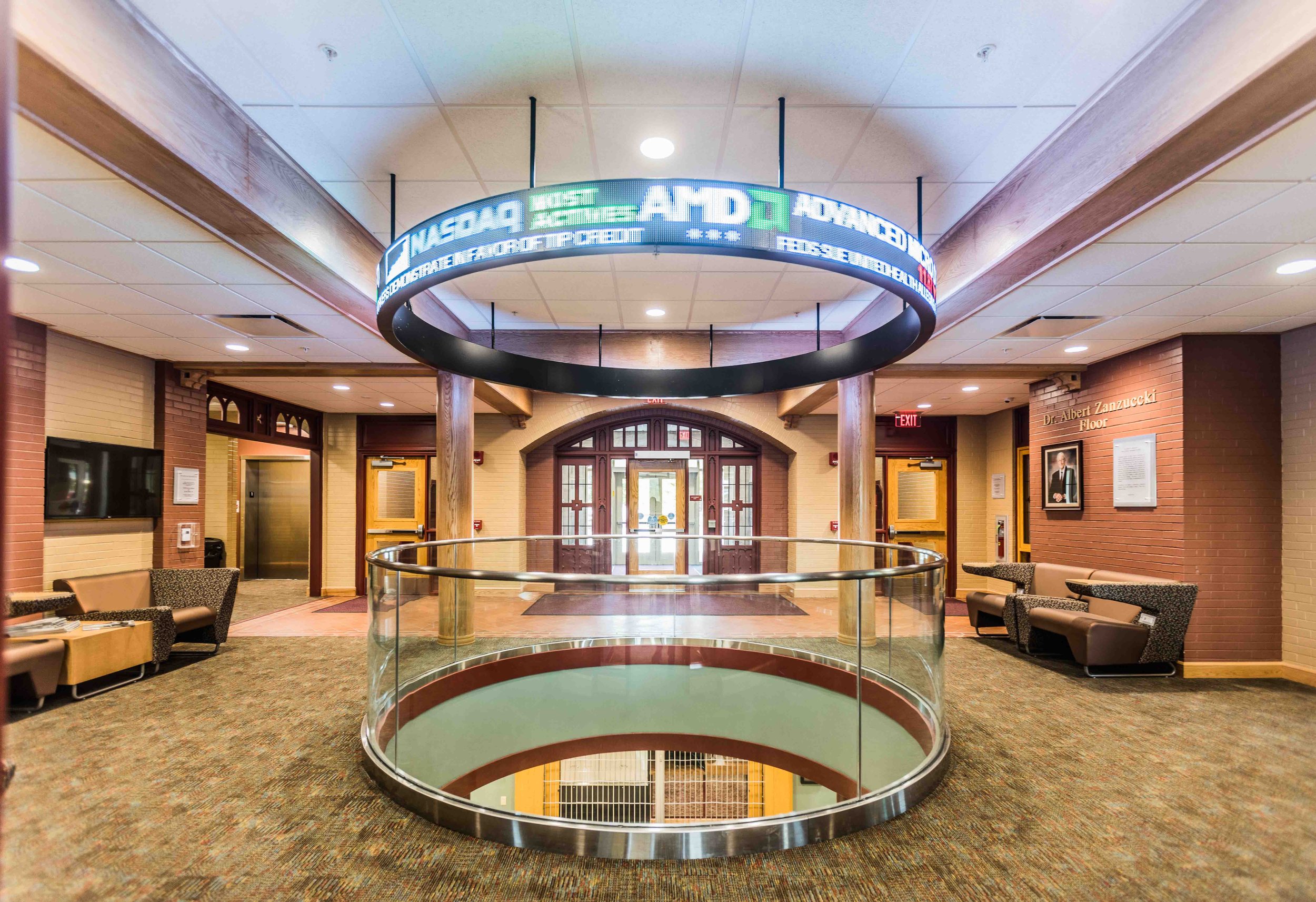 Inside a building with beige brick walls, carpeted floor, and a seating area with chairs. A circular opening in the floor with glass railing reveals stairs going down. Above, a digital ticker displays stock market information. Entrance doors are at t