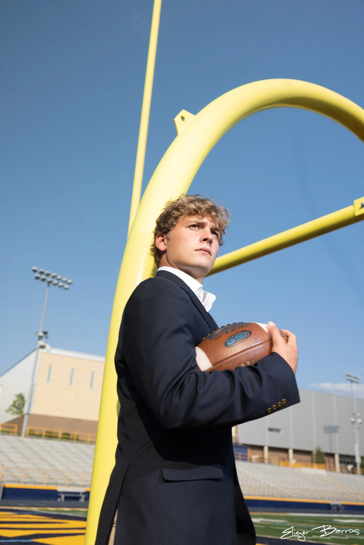 A young man in a suit holding a football, standing on a football field near a goalpost under a clear blue sky.
