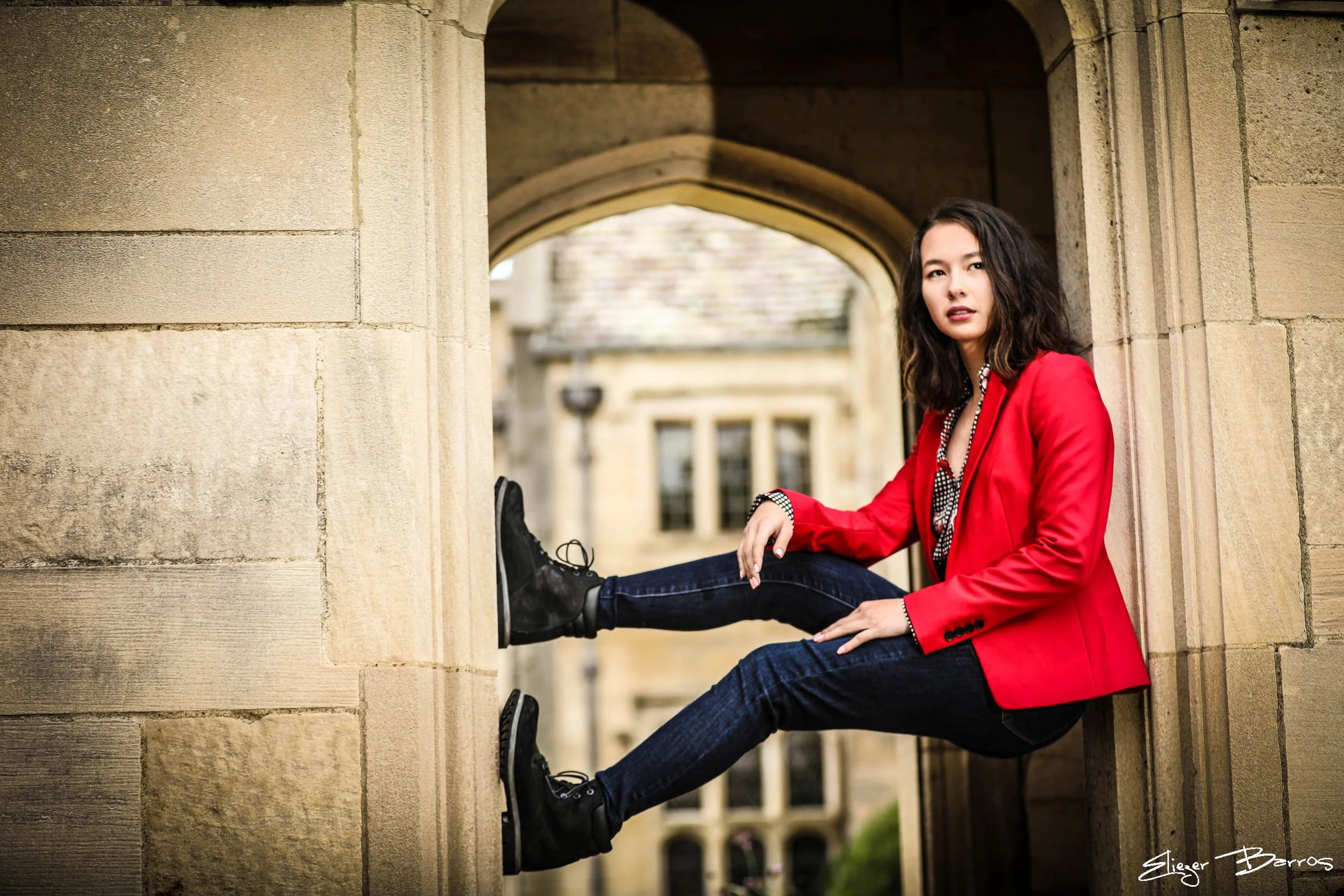 A woman sitting on a stone ledge under an archway, wearing a red blazer, black boots, and dark jeans, with her legs extended and crossed at the ankles, looking at the camera.