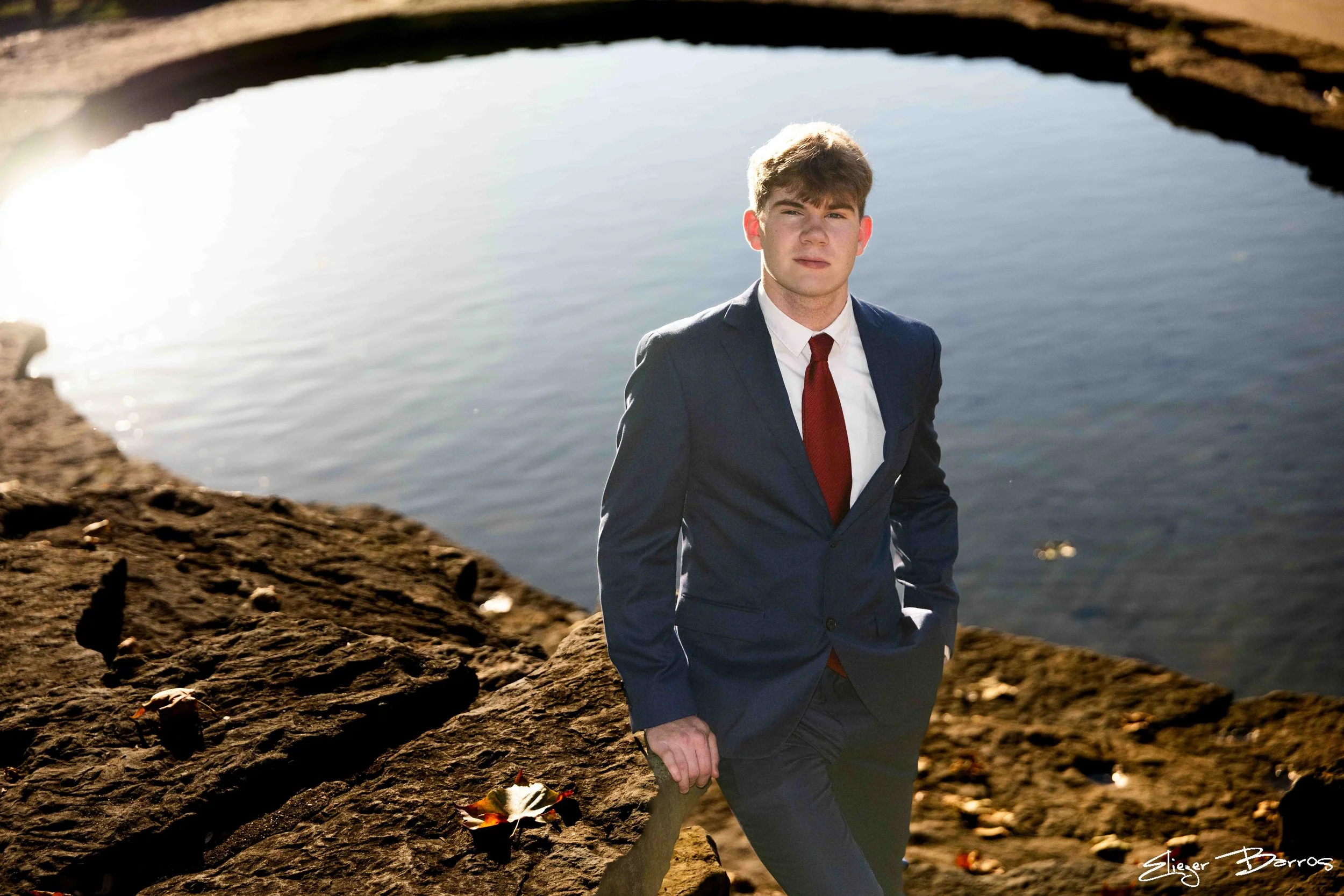 A young man dressed in a suit with a white shirt and red tie, standing outdoors near a body of water, with rocks and soil in the foreground and a reflection of the sky on the water's surface.