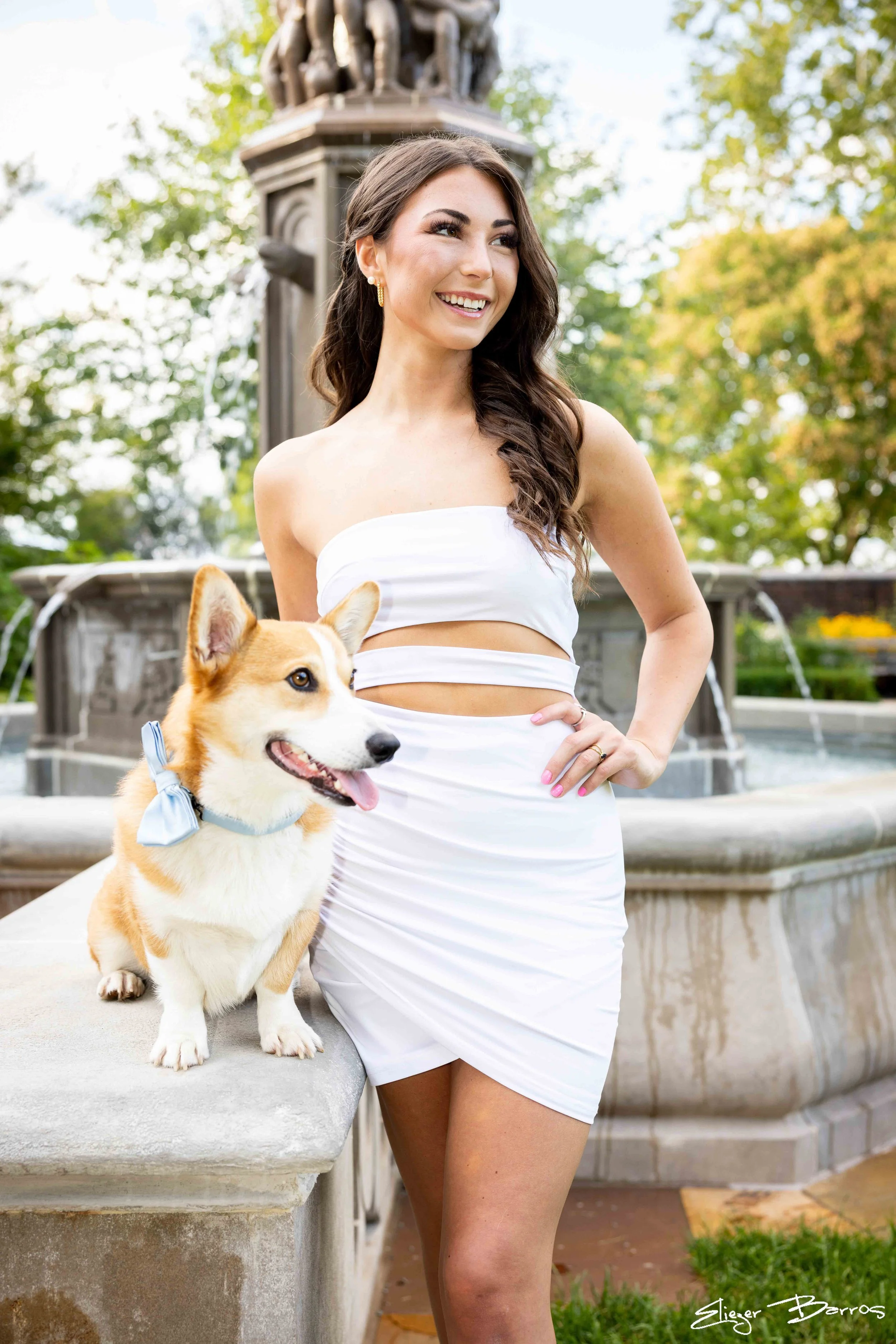 Senior Pictures Pittsburgh at Mellon Park. Young woman in a white strapless outfit posing with her corgi by a fountain during her stylish outdoor senior photoshoot.