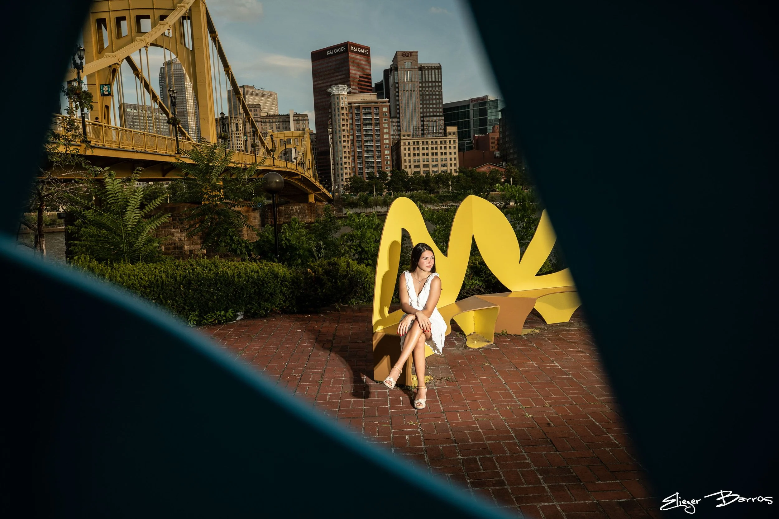 A woman in a white dress sitting on a yellow art installation at an outdoor location, with a city skyline including tall buildings and a bridge in the background, viewed through a dark frame.
